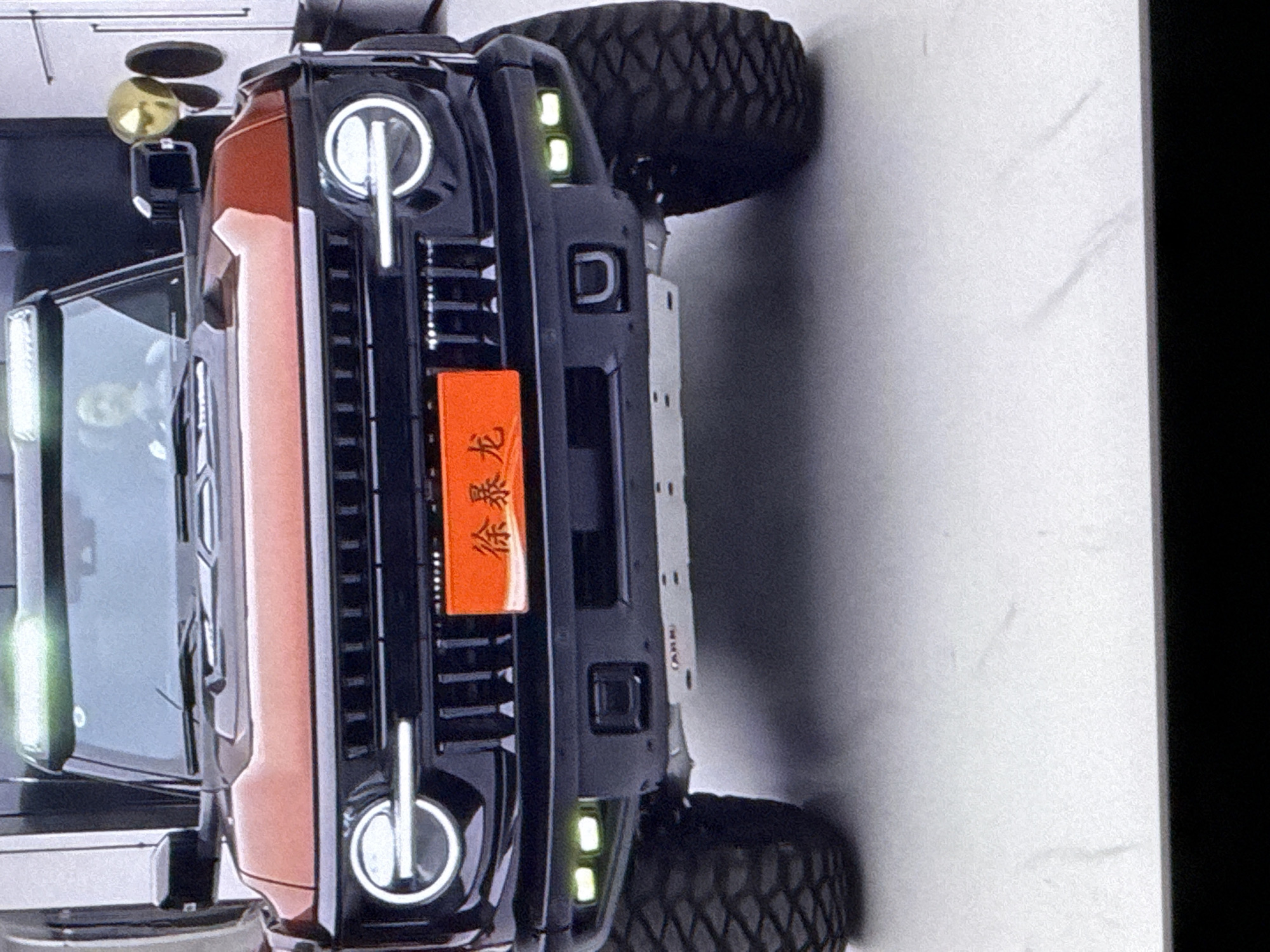 The image shows the front of an orange and black Ford Bronco SUV. The vehicle has a prominent black grille with vertical slats and circular headlights with LED accents. An orange license plate with Chinese characters is mounted on the grille. The SUV is equipped with auxiliary lights on the bumper and large, knobby tires. In the background, there's a person visible through the windshield, appearing to be the driver. To the right of the vehicle, a gold spherical object on a stand is visible, possibly a decorative item. The scene appears to be indoors, with a plain, light-colored floor and wall in the background, suggesting a showroom or garage. The image is underexposed in some areas, with a significant amount of digital noise. There is no indication of the city of Cahul, Moldova in the image.