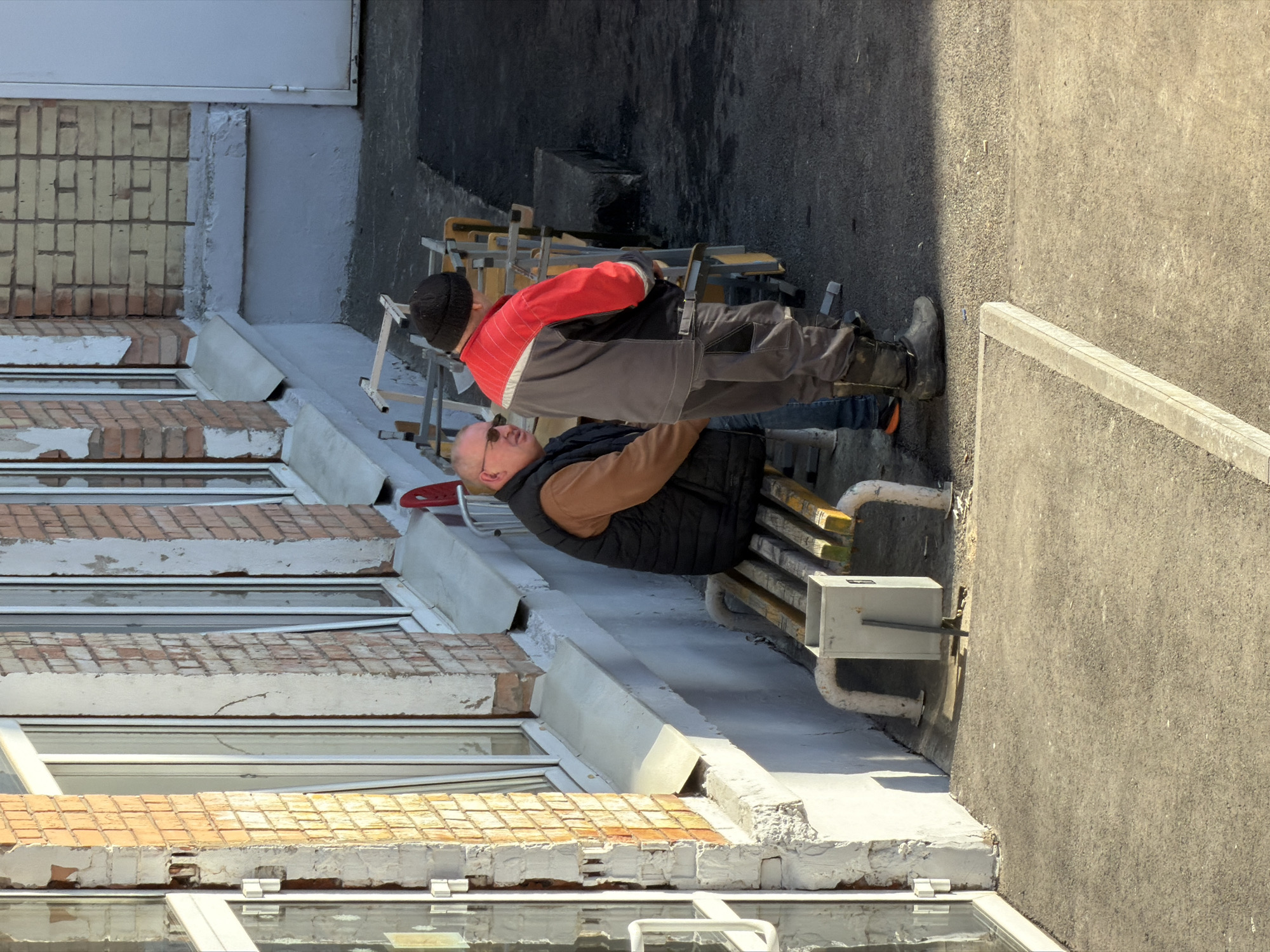 Two men are working on the exterior of a building. One man is wearing a black vest and brown shirt. The other man is wearing a gray jacket with red shoulders and black pants, as well as a black beanie. They are standing on a scaffolding platform next to a brick building with multiple windows. There are metal pipes running along the building exterior. The location is Izhevsk, Russia.