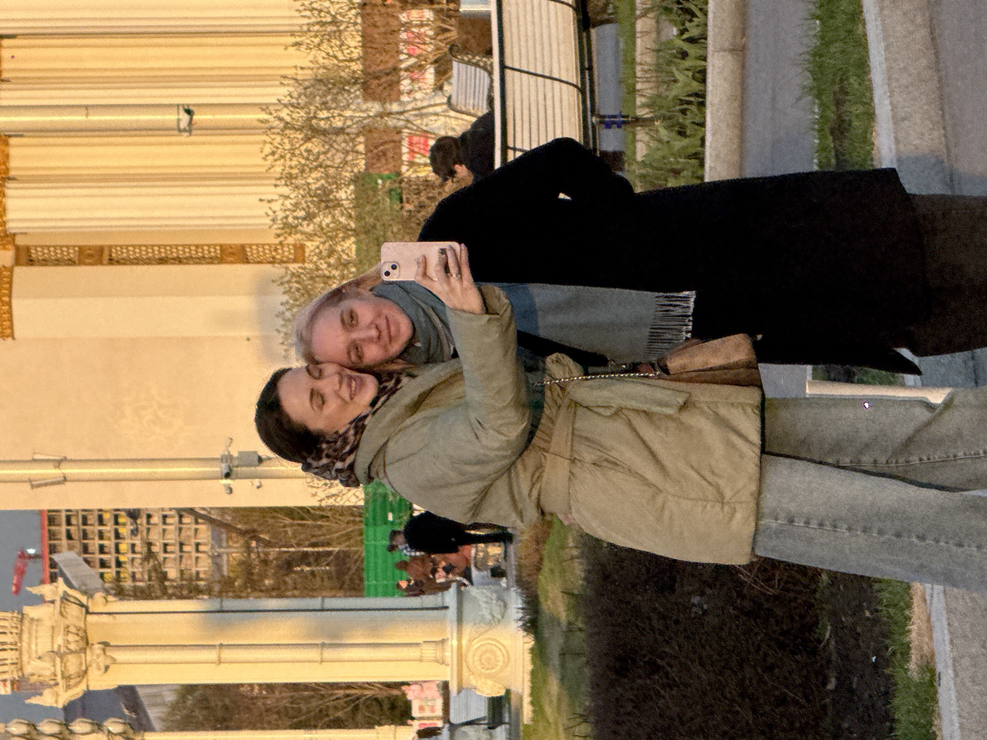 Two women are taking a selfie outside. The woman on the left has dark hair, wears a printed scarf, and a light brown coat. The woman on the right has light hair and wears a blue scarf and black coat. The location appears to be in Moscow, Russia, based on architectural elements in the background. They are standing on a sidewalk next to grass and bushes. Other people are walking in the background.