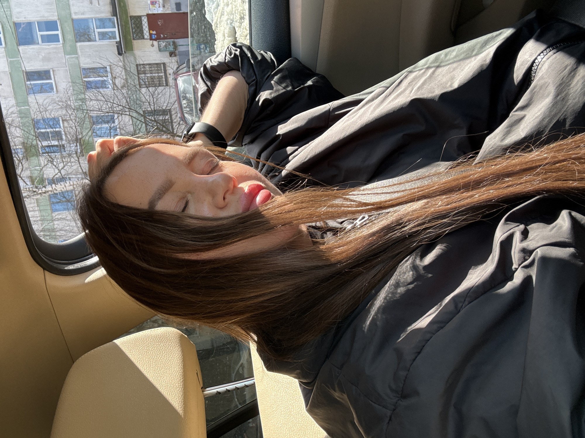 A woman with long brown hair lies in a car, presumably sleeping. Her face is tilted upwards, with her tongue slightly protruding from her mouth. She's wearing a black jacket. Outside the car window, there's a view of a multi-story building with numerous windows. This scene is located in the city of Chaikovsky, Russia.
