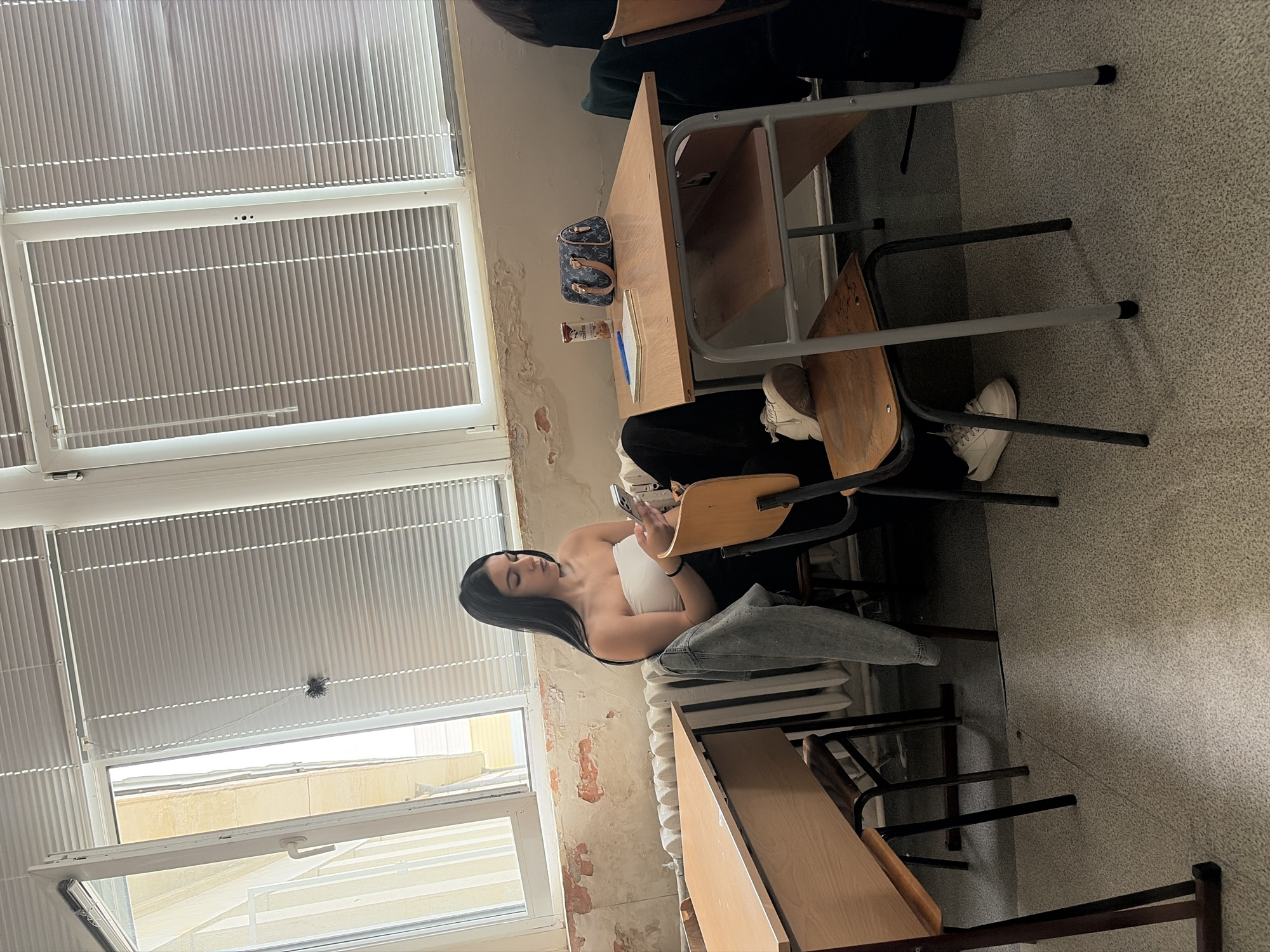 The image captures a young woman seated at a wooden desk in what appears to be a classroom or study area. She has long, straight dark hair and is wearing a white strapless top and light-colored jeans. Her gaze is directed downwards at a smartphone held in her right hand, suggesting she is engrossed in its content.
The setting features large windows on the left, covered by white vertical blinds that filter natural light into the room. Below the windows, a radiator is visible. The walls show signs of wear with peeling paint, revealing exposed plaster or brickwork underneath. The floor is covered with a light grey, speckled carpet.
On the desk in front of the woman, a small blue handbag adorned with the "Louis Vuitton" pattern is placed. Alongside it are a brown rectangular object, a carton of "Tedi" brand juice or milk, and a blue pen. The woman's feet, clad in white sneakers, are visible under the desk. Other wooden desks and metal-framed chairs are partially visible around her, contributing to the classroom ambiance.
The scene depicts a casual daytime moment, likely a break, with the woman occupying her time with her phone in an educational environment. The worn condition of the walls hints at an older building.