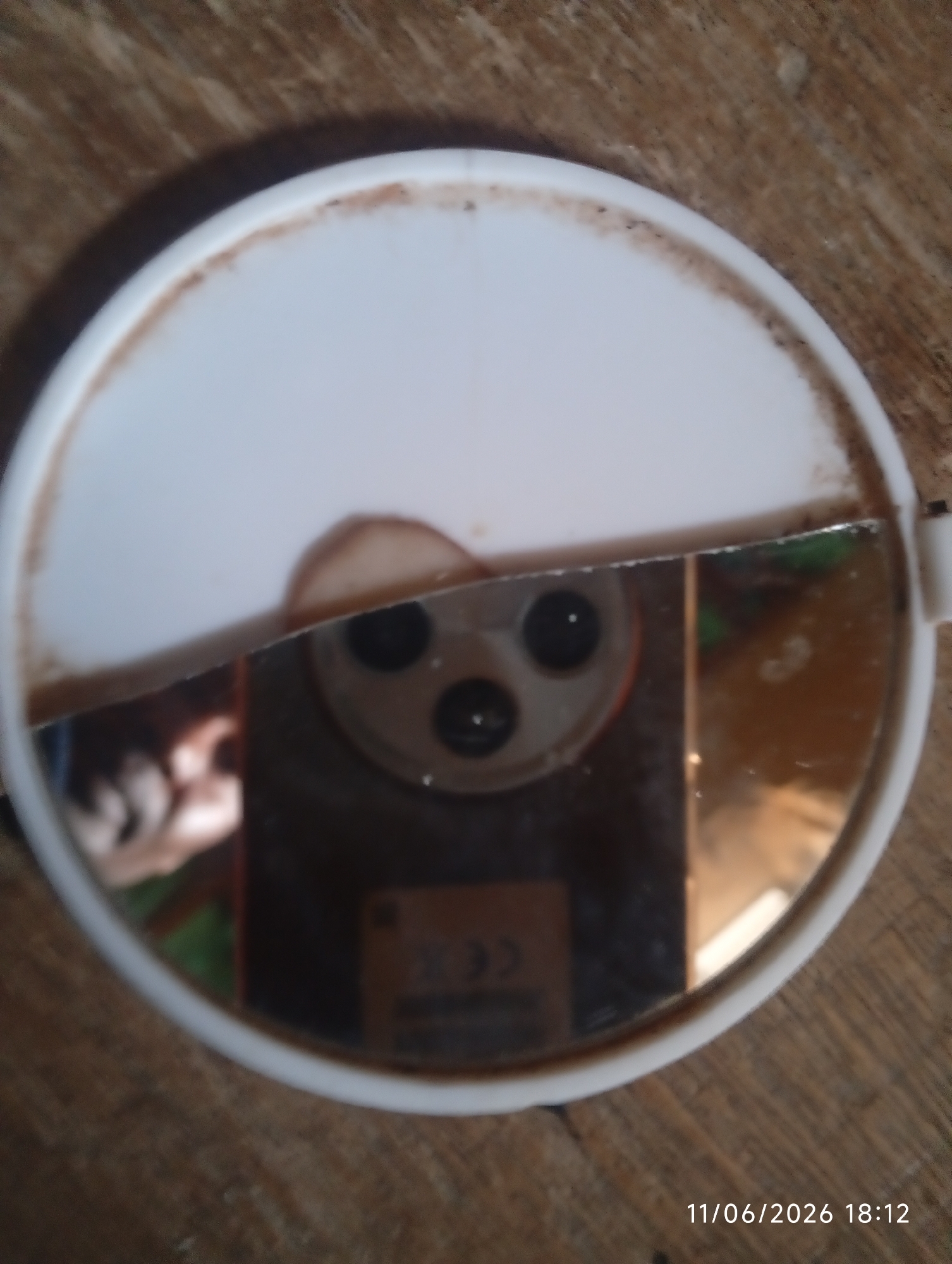 The image is a close-up, slightly blurred shot of a round, white-rimmed mirror with a cracked reflective surface. The mirror is resting on a wooden surface. The reflection in the mirror shows a person's hand and face, a camera lens system with three circular black lenses, and a portion of a smartphone screen with a partial view of a logo or text. The mirror is dirty around the rim. The timestamp "11/06/2026 18:12" is visible in the bottom right corner of the image. The location is not specified.
