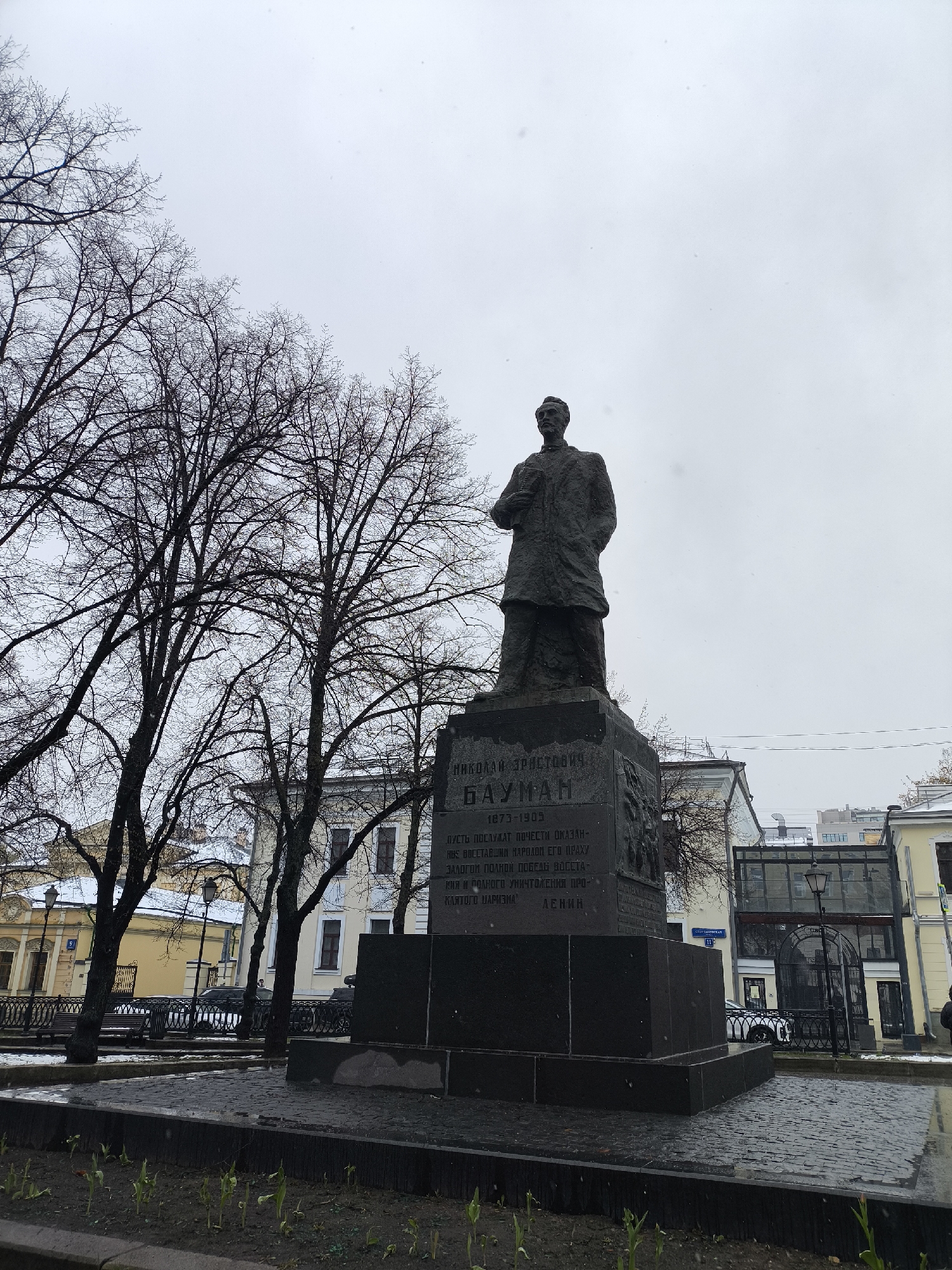 The media file presents a full-length statue of Nikolai Bauman, a prominent Russian revolutionary, situated in an urban park or square in Moscow, Russia. The dark-colored statue depicts Bauman in a long coat, his left hand seemingly holding documents, while his right hand is tucked into his jacket. He stands atop a multi-tiered, dark granite pedestal.
The pedestal bears Cyrillic inscriptions: "НИКОЛАЙ ЭРИСТОВИЧ. БАУМАН" and the years "1873-1905". Below this, a quote attributed to Lenin is inscribed: "ПУСТЬ ПОСЛУЖАТ ПОЧЕСТИ ОКАЗАН- НЫЕ ВОССТАВШИЙ НАРОЛОМ ЕГО ПРАХУ ЗАЛОГОЙ ПОЛНОЙ ПОБЕДЫ ВОССТА- КИЯ И ПОЛНОГО УНИЧТОЖЕНИЯ ПРО- КЛЯТОГО МАРИЗНА ЛЕНИН". Sculpted reliefs are also visible on the side of the pedestal.
The setting is characterized by numerous bare-branched trees, suggesting a late autumn or winter season. These trees frame the monument on the left and extend across the top of the image. The sky is uniformly overcast and light gray, contributing to a somber atmosphere. Small white specks are visible against the sky, possibly indicating light snowfall or fine precipitation.
In the background, a mix of architectural styles is visible, including older, light-colored buildings with dark window frames and a more modern structure featuring glass and dark metal. A black wrought-iron fence and a lamppost are also present. The ground immediately surrounding the monument is paved with dark cobblestones, and a narrow garden bed with emerging green shoots lines the base of the monument area. A white car is partially visible behind the fencing in the mid-distance. The scene conveys a quiet, historical urban landscape.