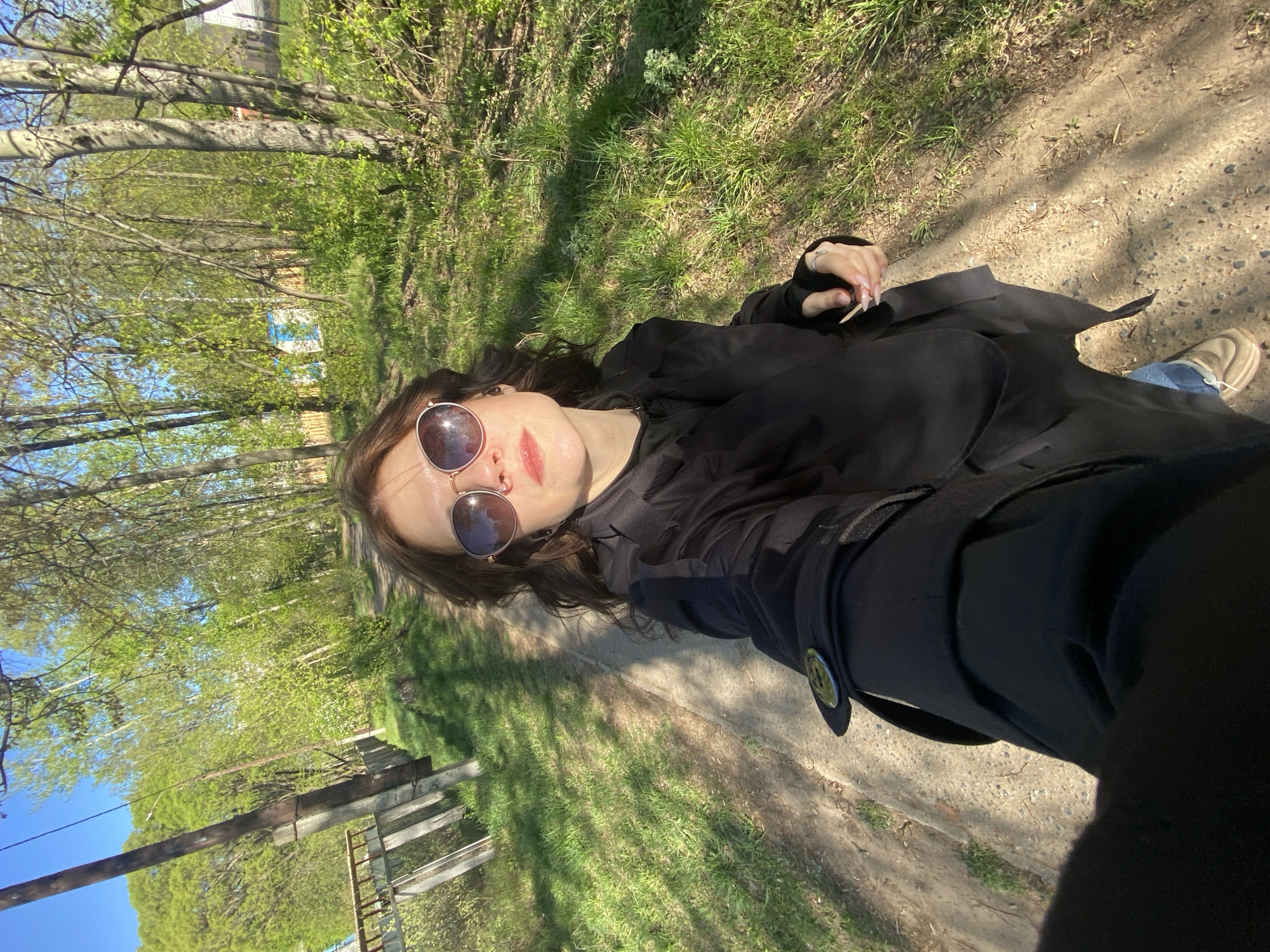 A woman in sunglasses and a black trench coat stands on a dirt path bordered by grass and trees. The scene is outdoors on a sunny day, likely in a park or wooded area of Chaikovsky, Russia. A wooden fence runs alongside the path, and residential buildings are visible in the background.