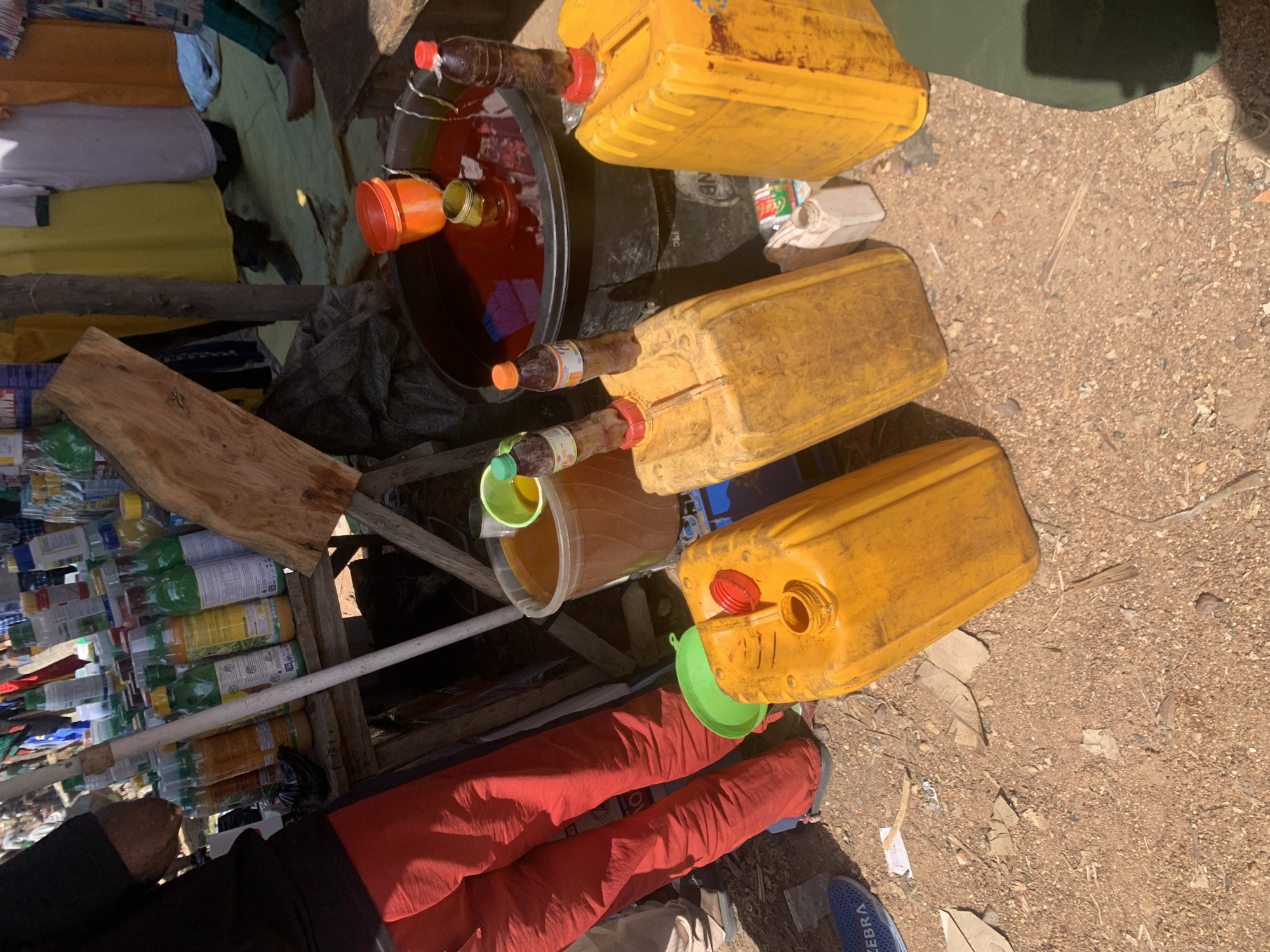 The image depicts an outdoor scene, likely a market stall in Nigeria, showcasing an array of items for sale, primarily liquids in various containers. The setting appears to be dusty and arid, with loose soil and pebbles covering the ground.
Dominating the frame are several large, yellow plastic jerrycans, some filled with a reddish-brown liquid, possibly palm oil or a similar commodity. One jerrycan has a bright red lid and another a green lid. Next to these, a large black basin holds a significant volume of the same reddish-brown liquid, with a few plastic bottles with orange and red caps submerged within it. Smaller plastic bottles, some appearing to contain beverages and others possibly oils, are stacked and displayed on a wooden shelf structure. These bottles vary in color and labeling, suggesting a range of products.
A piece of rough-hewn wood is positioned prominently in the foreground, possibly serving as part of the stall's display or construction. Partially visible in the lower portion of the image are the legs of at least one person, dressed in bright red trousers. Another person's hand, wearing a dark sleeve, is also visible. The overall impression is of a busy market environment where goods are being sold. The lighting suggests it is daytime, with harsh sunlight casting shadows. There is no visible text that is clear enough to be read. The scene conveys a sense of commerce and daily life within a Nigerian context.
