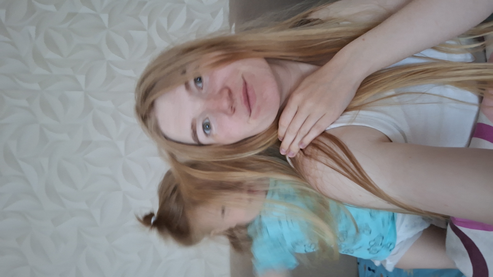 This photograph, taken in Chaikovsky, Russia, features an adult female and a young child relaxing together in an indoor setting. The image is oriented with the subjects lying on their side, looking towards the camera.
The adult, likely a mother, has long, light brown to blonde hair spread out around her head and shoulders. She has fair skin, blue-green eyes, and a gentle smile, indicating a relaxed and happy mood. She is wearing a simple white top, and her right hand rests near her neck.
Partially visible beneath her hair and to her side is a small child or infant. The child has light brown hair, tied in a small ponytail at the back of their head, and is wearing a light blue garment, possibly a t-shirt or part of a onesie, with faint patterned details. A white diaper is visible under the blue top. The child appears calm, lying close to the adult.
The background consists of a light-colored, possibly off-white or cream, wall with a distinct geometric or floral pattern, suggesting textured wallpaper. On the right side of the frame, there's a glimpse of patterned bedding or a blanket with white and pink/purple stripes, indicating they are likely on a bed or sofa. The lighting appears natural, suggesting it is daytime. The overall scene conveys a tender and intimate moment between the two individuals. There is no visible text in the image.
