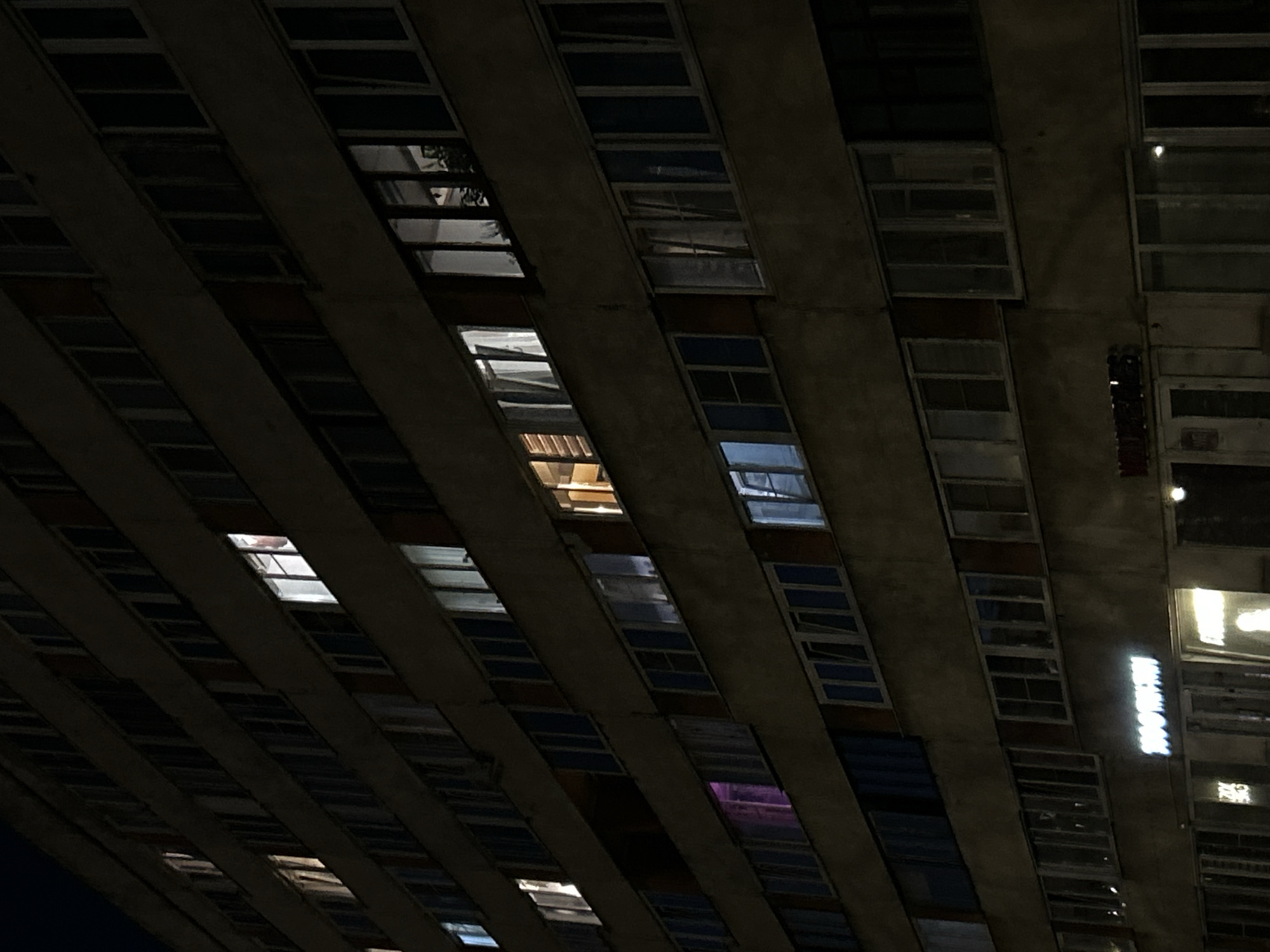 The image displays a multi-story building facade at night in Saint Petersburg, Russia. The structure is characterized by numerous rectangular windows arranged in long, diagonal rows, separated by dark concrete or stone bands. The architecture appears functional and repetitive, suggesting a large residential or commercial block.
The overall scene is dark, with the building's exterior largely unlit against the night sky. However, many windows glow with interior lights, creating a striking contrast. These lights vary in color, with some casting warm yellow hues, others cool white, and a few emitting a distinct purple glow. Some windows are completely dark, while others offer reflections of unseen elements or glimpses into dimly lit interiors.
No people are visible, and no specific activity is overtly depicted on the exterior. The illuminated windows imply ongoing life and activity within the building, fostering a sense of urban anonymity and quietude typical of a city after dark. A notable detail is bright, illuminated text visible through a window on the right side of the building. This text, appearing in white Cyrillic script, is discernibly "МАГНИТ" (MAGNIT), suggesting the presence of a commercial establishment, likely a supermarket, on one of the lower levels. The clear reflections seen in some window panes suggest calm weather conditions.