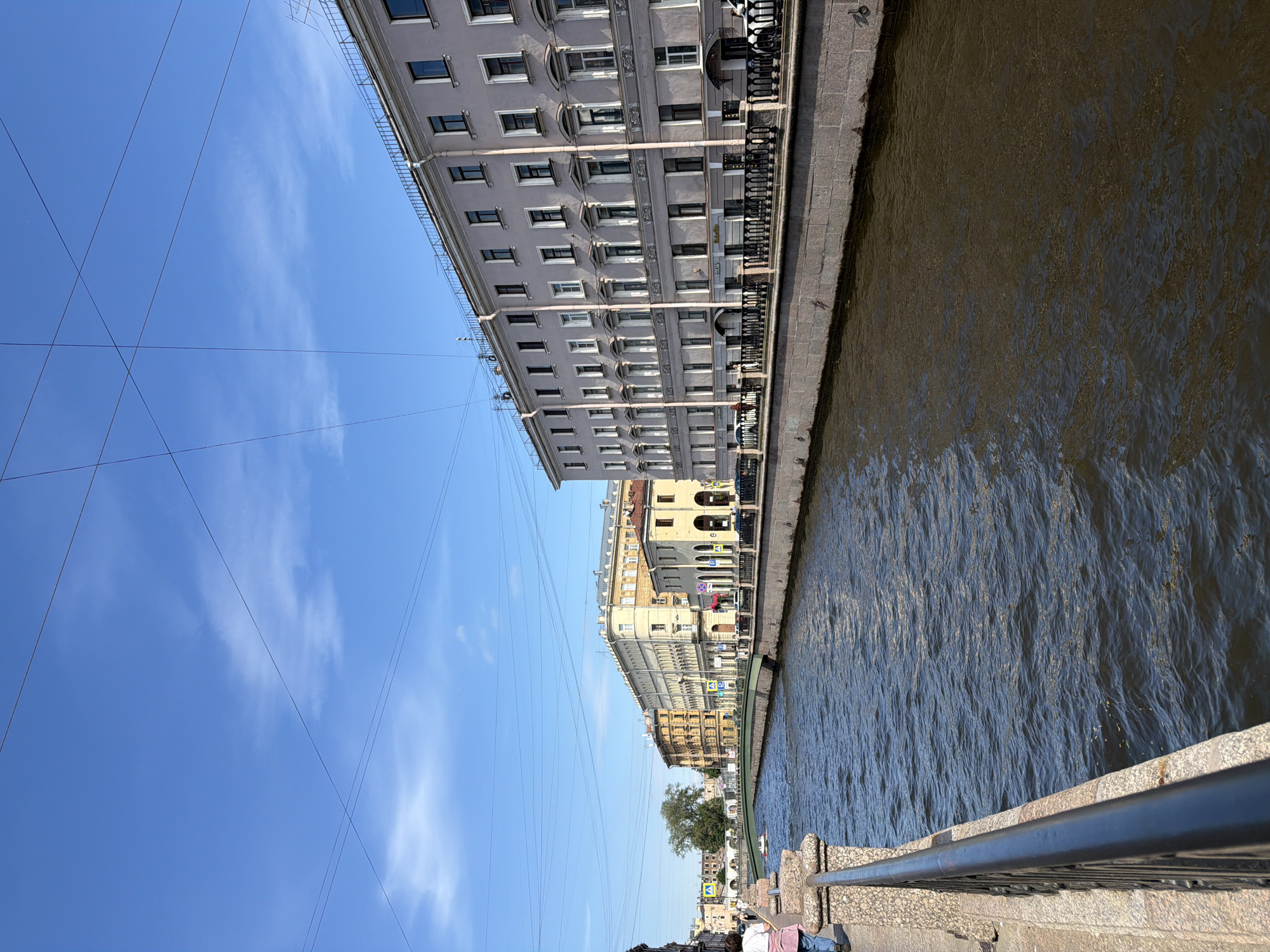 The image shows a wide canal with buildings lining its banks. A green bridge spans the canal in the distance. The water is a dark, rippling color, reflecting the bright blue sky and white clouds. The buildings on the right are tall and appear to be multi-story residential or commercial structures with many windows. They have a light gray facade and ornate architectural details. A black wrought-iron fence runs along the top of the canal wall. On the left bank, a person in a pink jacket and blue jeans is walking away from the camera, and another person is visible further down the embankment. The canal is a prominent feature, suggesting it is a waterway within a city. Considering the architectural style and the presence of canals and bridges, the scene is likely Saint Petersburg, Russia. The overall atmosphere is one of a clear, sunny day in an urban environment.