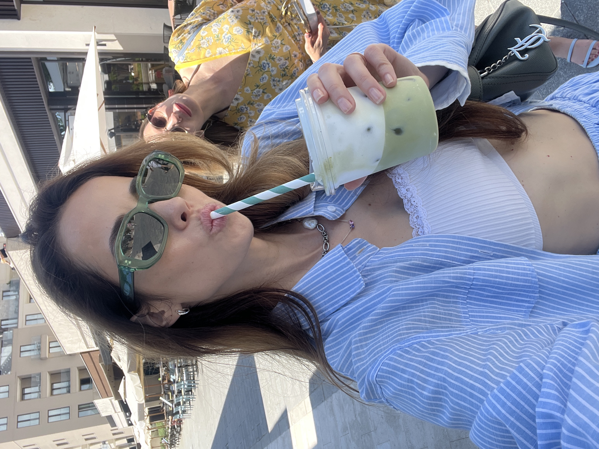 This image is a selfie taken by a woman outdoors, capturing herself and another woman in the background. The primary subject, in the foreground, has long brown hair and wears green sunglasses. She is puckering her lips around a green and white striped straw, drinking from a clear plastic cup containing a layered green and white beverage, possibly a matcha latte. She is dressed in a light blue and white striped oversized shirt, worn over a white lace bralette or top, and a silver chain necklace with a large pearl pendant is visible around her neck.
Partially visible in the background is another woman, out of focus, wearing a yellow floral dress and sunglasses. A dark green handbag with a white "Juicy" keychain hangs near the foreground woman's shoulder, and light blue sandals are visible on the background woman's foot.
The scene is an outdoor setting, likely a cafe or a recreational area in Tivat, Montenegro, with modern-looking buildings, white awnings, and paved ground in the background. Bright sunlight casts distinct shadows, indicating a sunny day, possibly midday or early afternoon. The overall atmosphere is relaxed and pleasant, suggesting a leisurely moment between the two women.