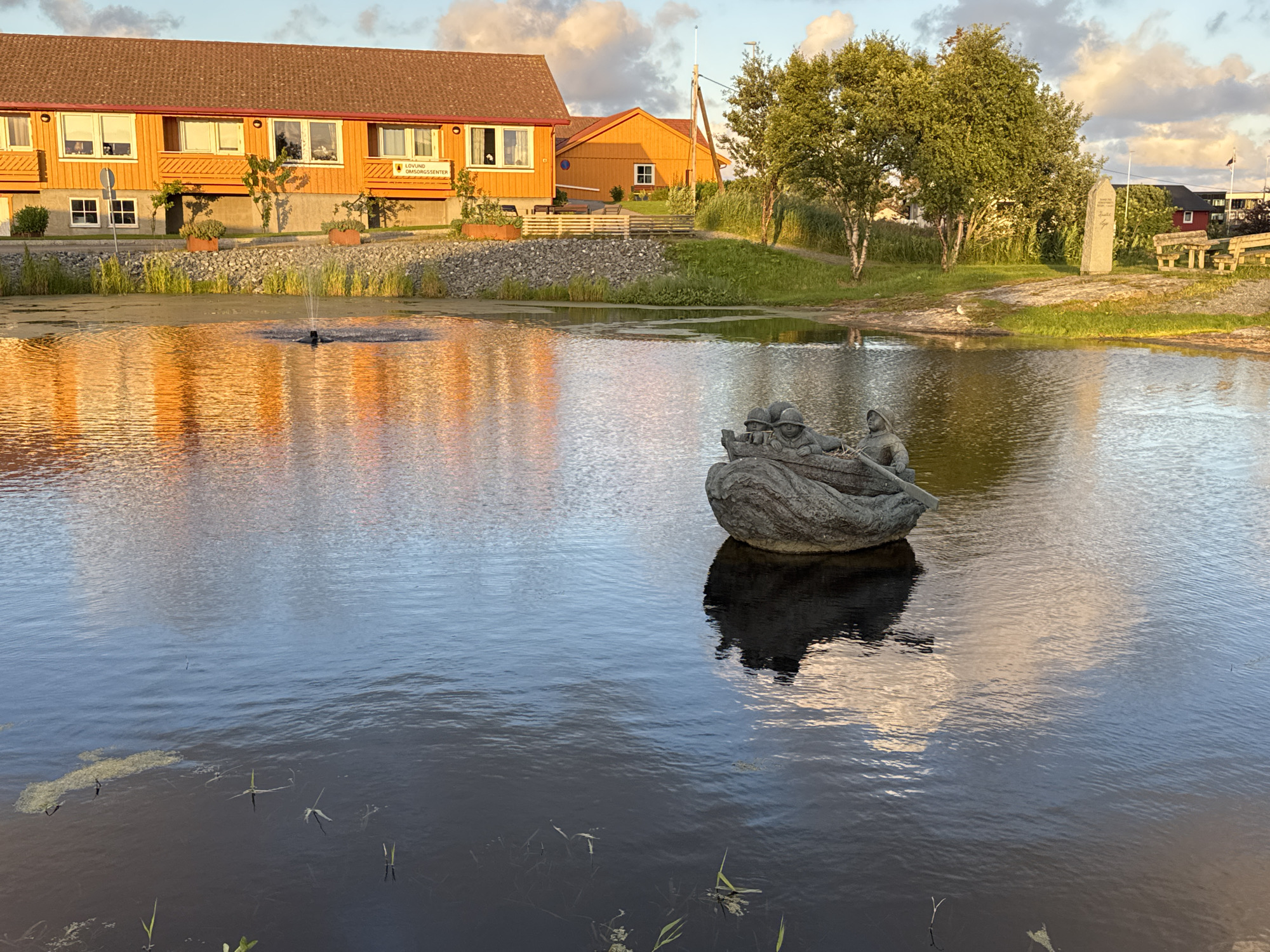 In Lovund, Norway, a scenic view unfolds with a golden-hued building reflecting in a calm body of water. The building, a long, two-story structure with a reddish-brown roof, features multiple windows and a sign reading "LOVUND OMSORGSSENTER". A small fountain spouts water in the foreground, creating ripples that distort the reflection of the building and the surrounding greenery. In the water, a stone sculpture depicts figures in a boat, with its shadow cast onto the pond's surface. To the right of the water, a grassy embankment is dotted with trees and a tall, slender stone monument. In the background, more buildings and trees are visible under a sky with scattered clouds.