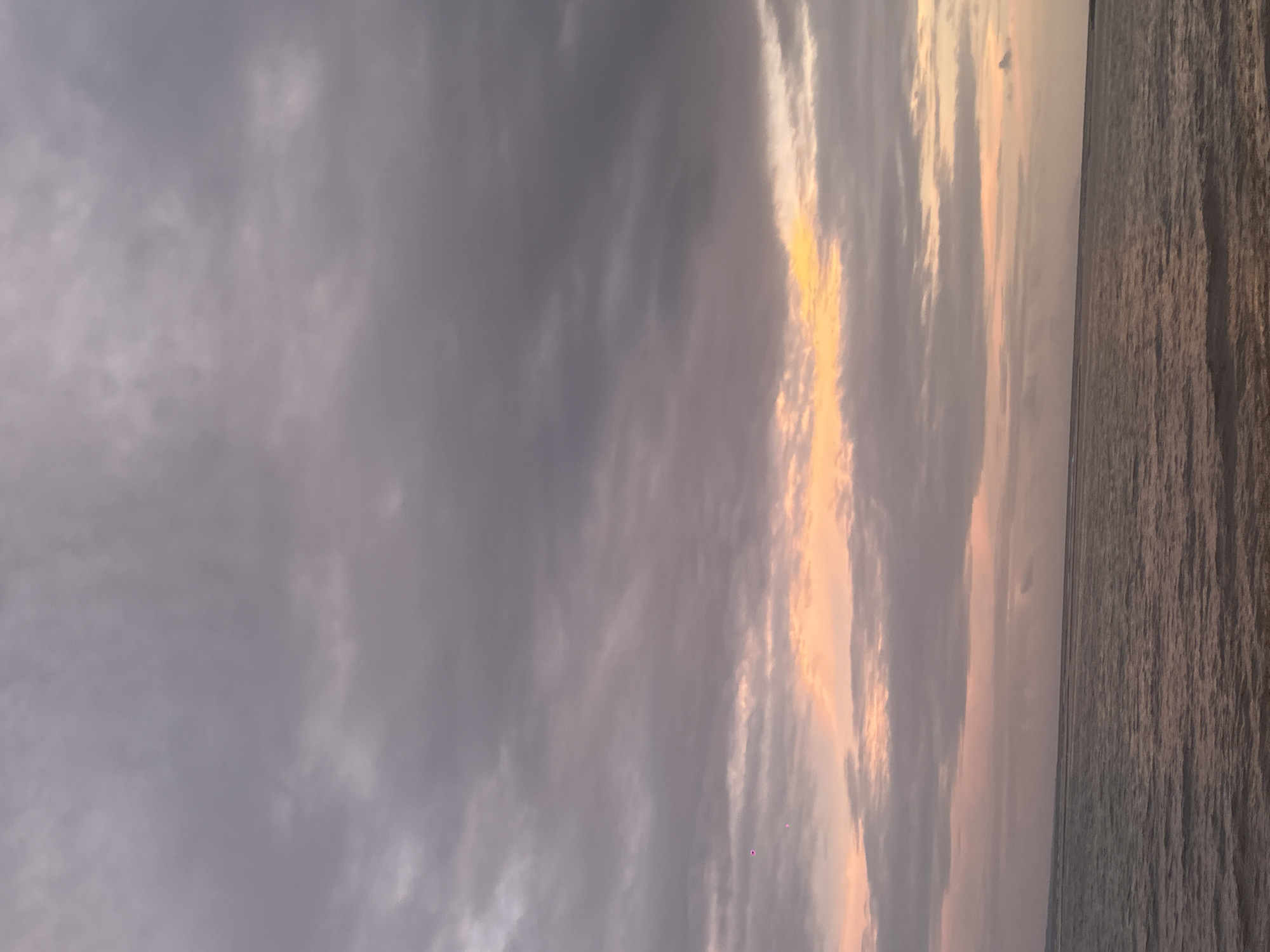 The image captures a landscape scene at dusk or dawn. The right side of the frame is dominated by a body of water, possibly the sea, with its surface reflecting muted colors of light. The left side of the image is primarily the sky, filled with layers of grey and white clouds. There is a patch of golden light breaking through the clouds, suggesting the presence of the sun obscured by the overcast. The composition is somewhat simple, with the horizon line appearing relatively straight, separating the water from the sky. There are no visible people or objects in the foreground or background. It is not possible to verify that this scene is specifically in the city of Sainte-Anne, France, with no further information.