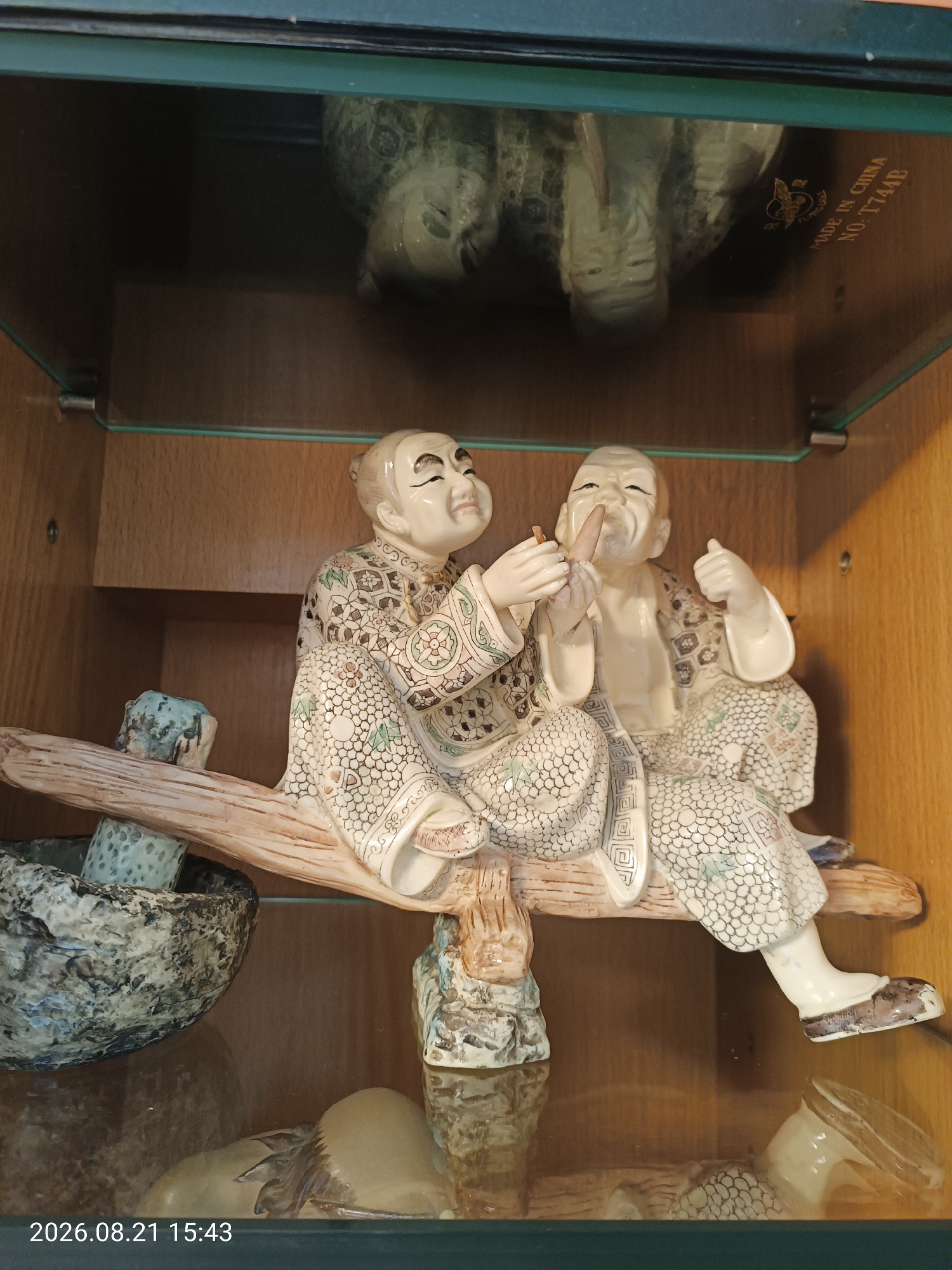 An antique ivory-colored figurine of two elderly East Asian men, each wearing patterned robes and seated on a wooden branch, is displayed within a glass cabinet. The man on the left holds a small brown object to his mouth, while the man on the right gives a thumbs-up gesture. A carved stone bowl sits to the left of the figurine. A label on the glass cabinet reads "MADE IN CHINA NO. T744B". The timestamp "2026.08.21 15:43" is visible in the lower left corner of the image. The scene is likely inside a private residence or museum in Hong Kong, China.