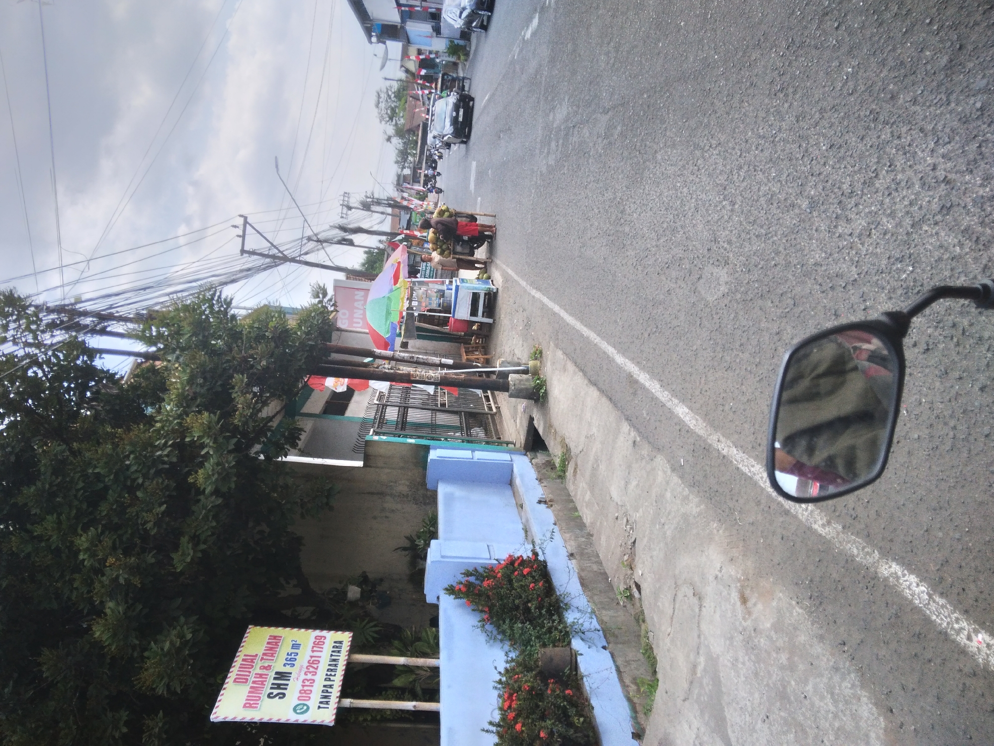 The image depicts a street scene in Sokaraja, Indonesia, characterized by a roadside with a blue-painted wall and a sidewalk. A prominent yellow sign on the left advertises "DIJUAL RUMAH & TANAH SHM 365 m²" with contact information. Trees with dense foliage are visible behind the sign. Further down the street, a colorful umbrella shades a small stall where two individuals appear to be engaged in commerce, possibly selling fruit, with a cart laden with produce nearby. Several motorcycles and cars are present on the asphalt road, indicating moderate traffic. Power lines crisscross the sky, and on the right side of the road, buildings and more flags suggest a commercial or residential area. The presence of the yellow sign and the general street layout are consistent with typical urban environments in Sokaraja.