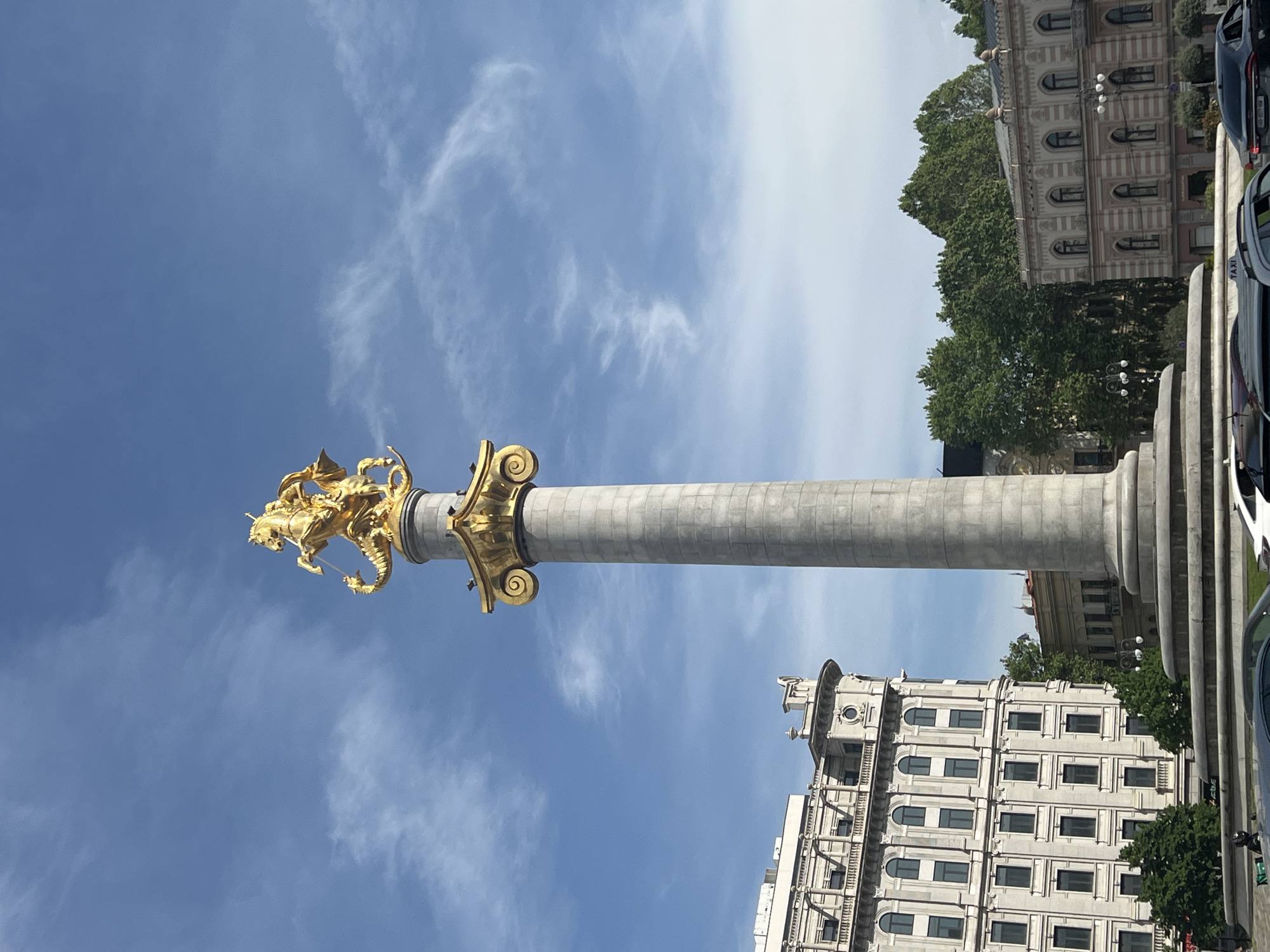 The image shows the St. George Statue atop the Pillar of Freedom in Tbilisi, Georgia. The statue depicts St. George slaying a dragon. The pillar is light gray with a gold Corinthian capital. The sky is blue with scattered white clouds. Buildings with light-colored facades are visible in the background, along with some trees and cars. The perspective is a low angle looking up at the monument.