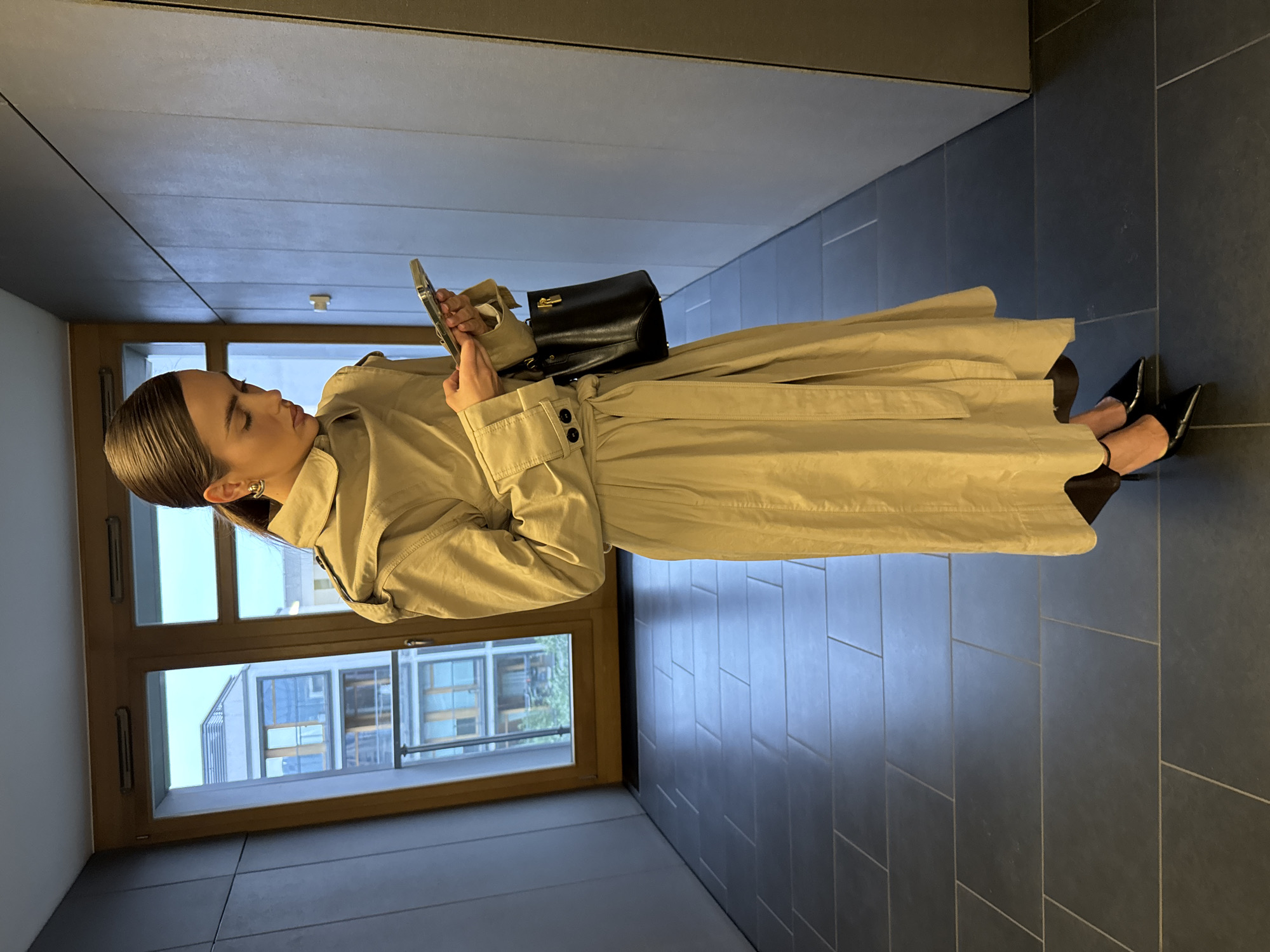 A woman stands in a hallway with dark gray tile flooring. She is wearing a tan trench coat, black heels, and carrying a black handbag. She is looking at her phone. Behind her is a window showing a view of buildings. The location is a hallway in a building in an unknown city.