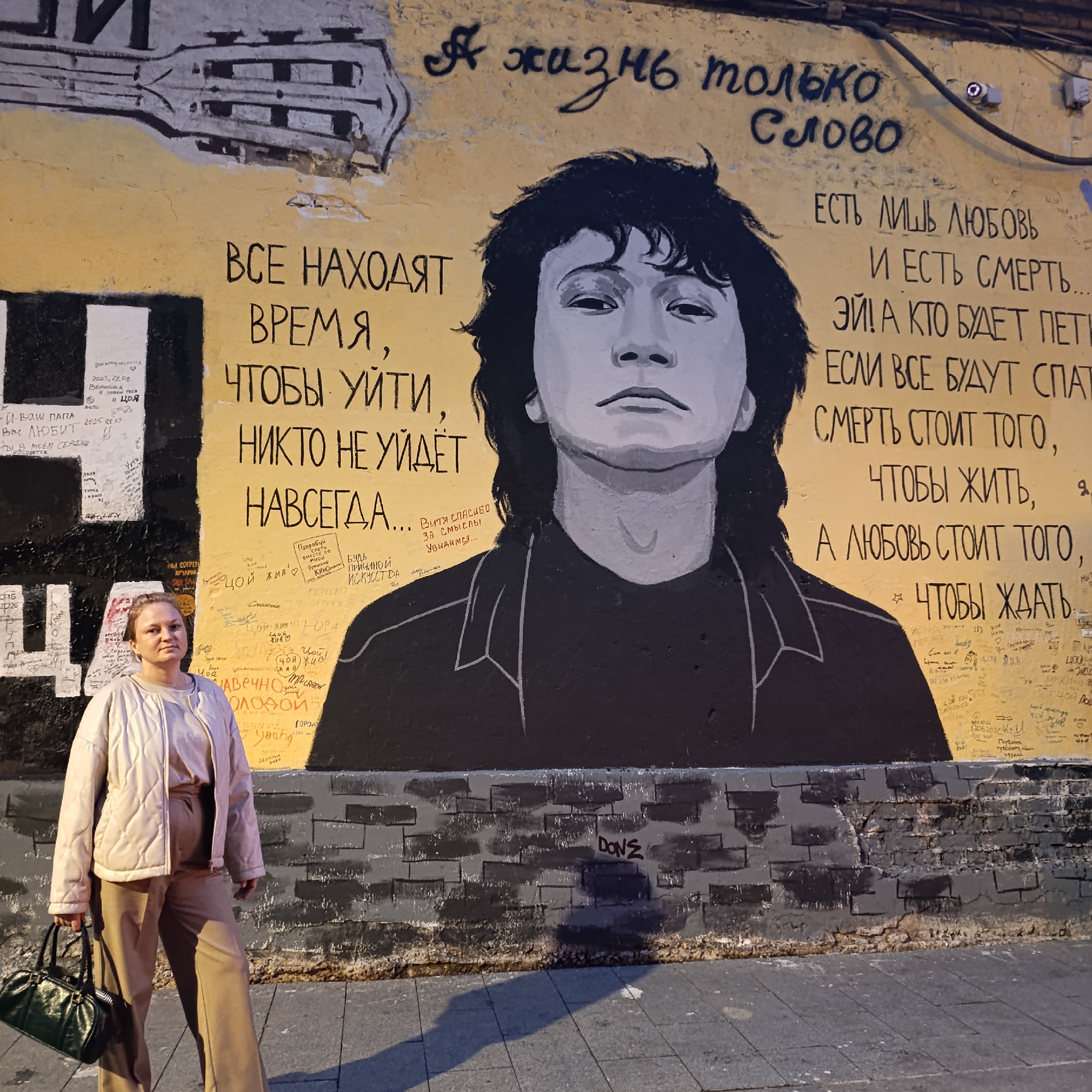 The image shows a mural of Viktor Tsoi on a yellow wall, located in Moscow, Russia, on Arbat Street. The mural depicts Tsoi's face in black and white, with a guitar motif above him. To the right of the mural, there is Russian text that translates to "And life is only a word." Below this, more text reads "There is only love and there is death... so who will sing... if everyone will sleep, death is worth it, to live, and love is worth it, to wait." There are also numerous smaller inscriptions and graffiti covering the wall. A woman stands in front of the mural, wearing a quilted jacket and trousers, holding a dark green handbag. The mural is situated on a brick wall that is painted black in sections, with a sidewalk in front of it.