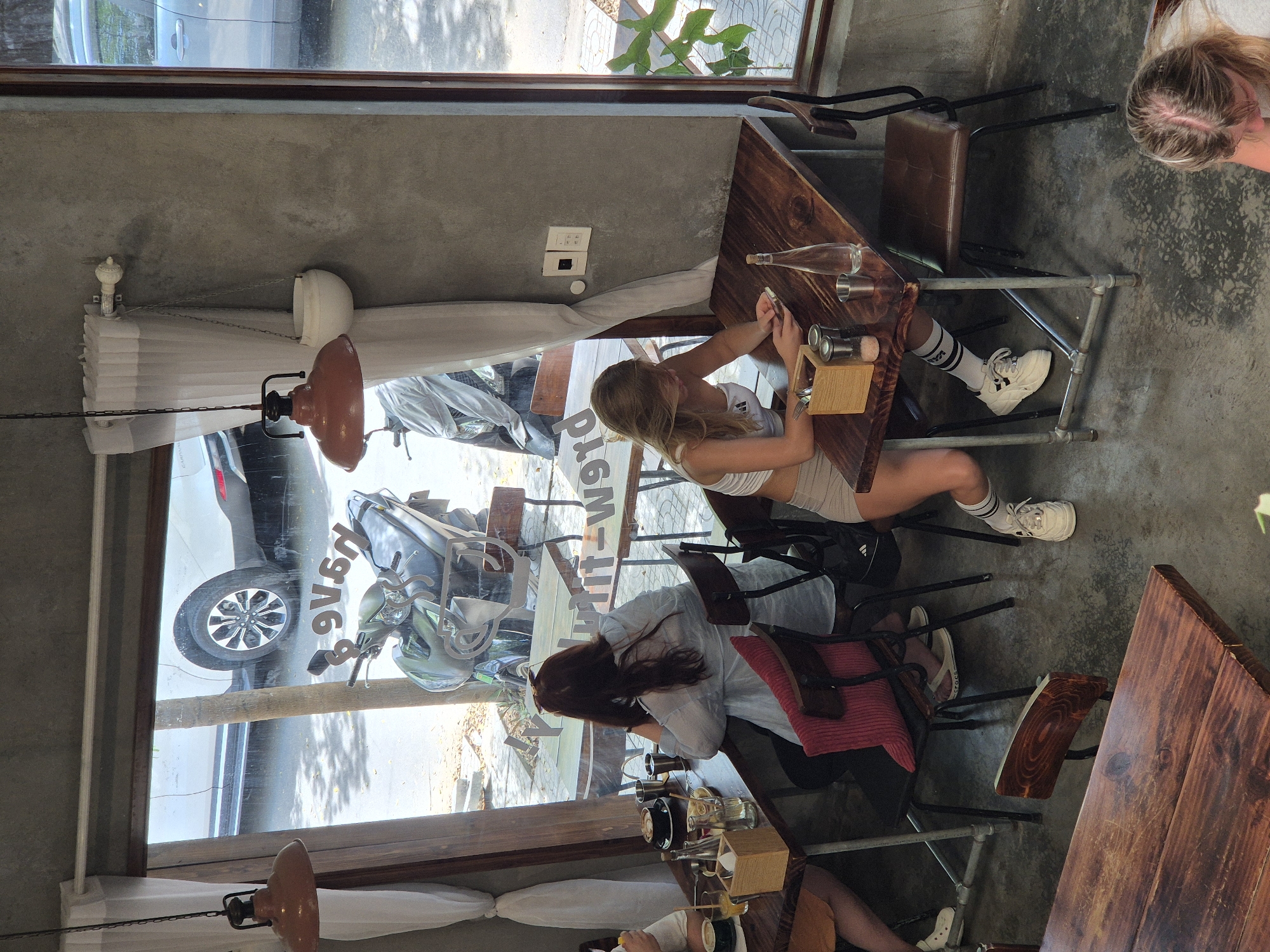 The image depicts the interior of a modern cafe in Da Nang, Vietnam, with a large window overlooking a street. Inside, two women are seated at separate tables. One woman, with long dark hair, is seated at a table to the left, looking towards the window and resting her chin on her hand. She is wearing a light blue shirt and has a pink cushion on her chair. On her table, there is a coffee cup, a small plate with food, glasses, and a wooden box with utensils.
The second woman is seated at a table on the right, facing the camera and looking down at her phone. She has blonde hair and is wearing a white crop top and light-colored shorts with striped socks and white sneakers. On her table are salt and pepper shakers, a wooden utensil holder, a glass bottle, and a small metal cup.
The cafe features concrete walls and wooden tables and chairs. Outside the window, a white car and motorcycles are visible on the street, along with trees casting shadows on the ground. The cafe is decorated with hanging lamps and a white pot suspended from the ceiling. The overall atmosphere suggests a casual dining or coffee shop environment.