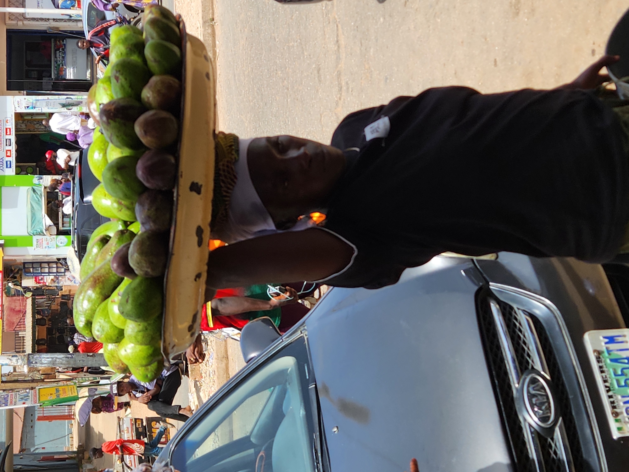 A medium shot depicts a woman balancing a tray of avocados and passion fruit on her head next to a parked Kia car. The woman, wearing a black shirt with white trim, a white head covering, and a yellow patterned turban underneath, looks toward the camera. The Kia car, with a visible license plate, is parked on a street with a clear sky overhead. The background features urban elements, including storefronts, people, and other cars, all suggesting a bustling market or commercial area. The location is specified as Unknown, Unknown.