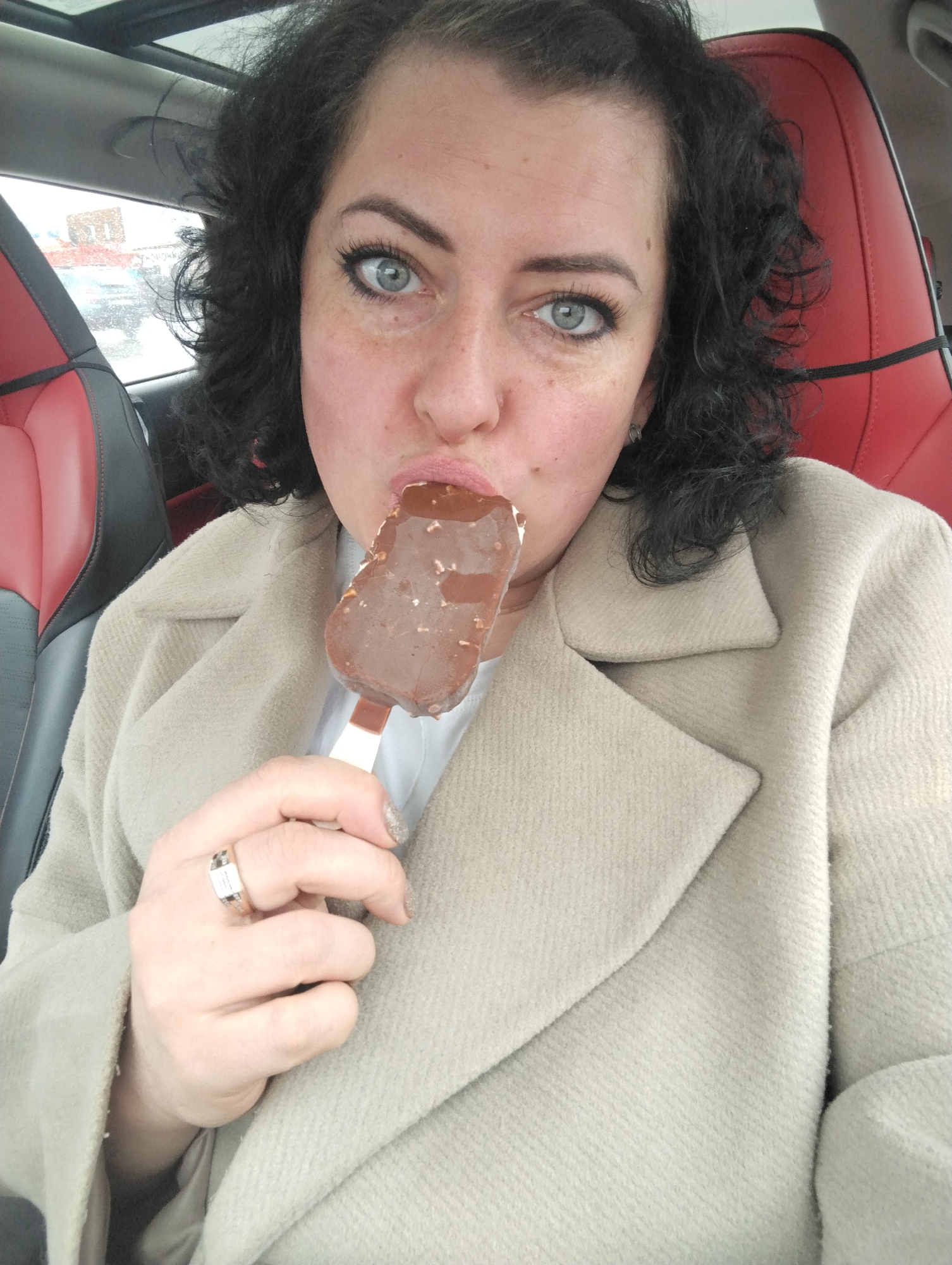 A woman is captured in a close-up selfie inside a car, holding and eating a chocolate-covered ice cream bar. She has dark, curly hair, blue eyes, and is looking directly at the camera with an open-mouthed expression as if about to take a bite or having just taken one. Her lips are slightly parted, touching the melting ice cream. She is wearing a light-colored (beige or cream) long coat over a white top. A silver-colored ring with a rectangular face is visible on her right ring finger, and her nails have a glittery polish.
The setting is the interior of a vehicle, suggested by the red and black leather car seat visible behind her, as well as the dark ceiling and a partial view of a sunroof. Through the car windows in the background, a blurred outdoor scene suggests a cloudy or overcast day, possibly with precipitation, implying the location is Izhevsk, Russia, as per the provided context. The activity central to the image is the woman enjoying the ice cream, which has visible nuts or crunchy bits embedded in its chocolate coating. No discernible text is visible in the image.