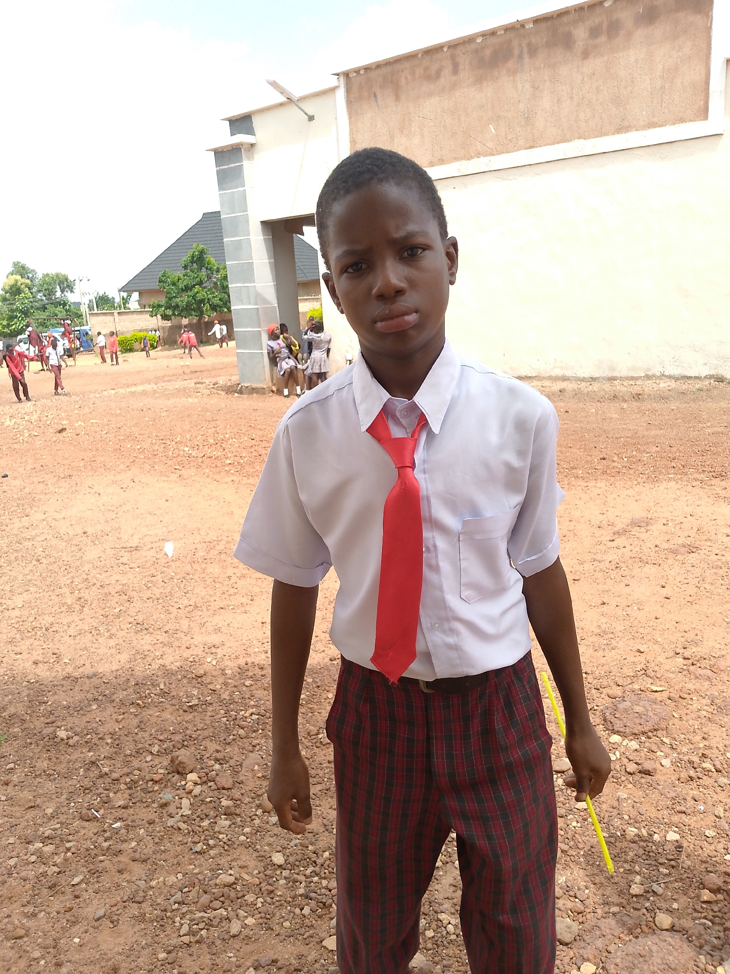A young boy, appearing to be of school age, stands in a dusty outdoor area. He is wearing a white short-sleeved shirt, a red tie, and plaid pants. His left hand is at his side, and he holds a yellow stick in his right hand. In the background, other children are visible, some wearing similar school uniforms. There is a building with light-colored walls and a dark-tiled roof behind the children. The ground is covered in dirt and small stones. The sky is overcast. The image appears to have been taken at ground level.