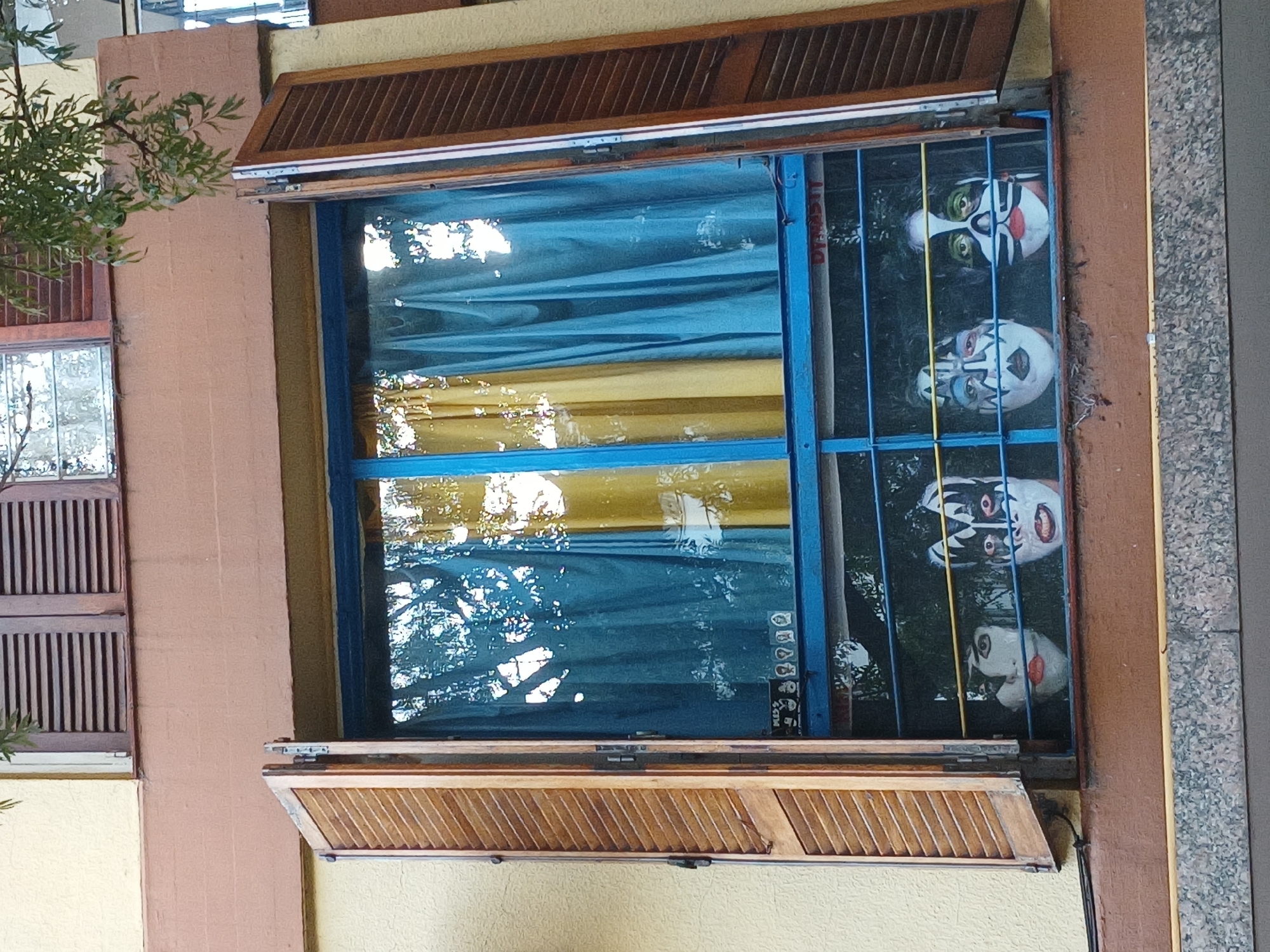 A photograph depicts a building facade with two windows. The larger window, framed in blue with a protective metal grill, displays four KISS-themed masks lined vertically. The window has wooden louvered shutters, with the upper and lower sections open, revealing a blue and yellow curtain within. The smaller adjacent window, similarly fitted with wooden louvered shutters, is partially visible on the left side of the frame. The building exterior is painted in a combination of yellow and brown tones. The location is likely in the city of Buenos Aires, Argentina, as indicated by the Dynastit logo on the window frame. The masks are positioned behind the bars of the window's protective grill.