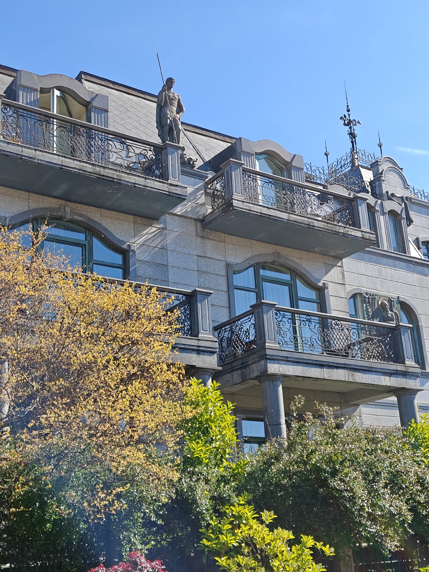 The image depicts the facade of a multi-story stone building in Batumi, Georgia, characterized by architectural elements such as balconies with wrought-iron railings, arched windows with green frames, and a slate-tiled roof. A statue of a man holding a spear stands atop the building. In the foreground, there are trees with yellow blossoms and green bushes, partially obscuring the lower levels of the building. The sky is clear and blue. The architecture and details suggest a design influenced by European styles.