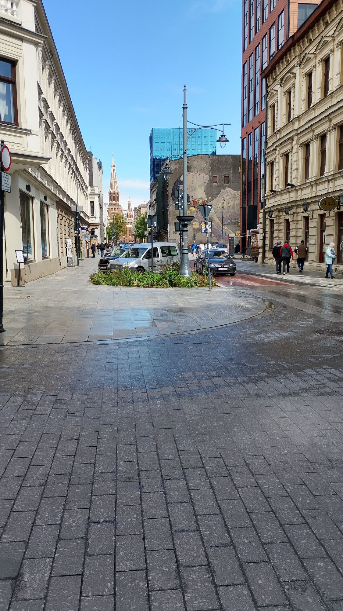 The image depicts a wet, pedestrianized street in Łódź, Poland, paved with gray rectangular blocks. A circular island with vegetation and a lamppost is located near the center of the image. Two cars are parked beside the island. Several people are walking on the sidewalks along the buildings on either side of the street. A red brick church tower is visible in the distance down the street. A tall blue glass building and a mural on a building can also be seen.