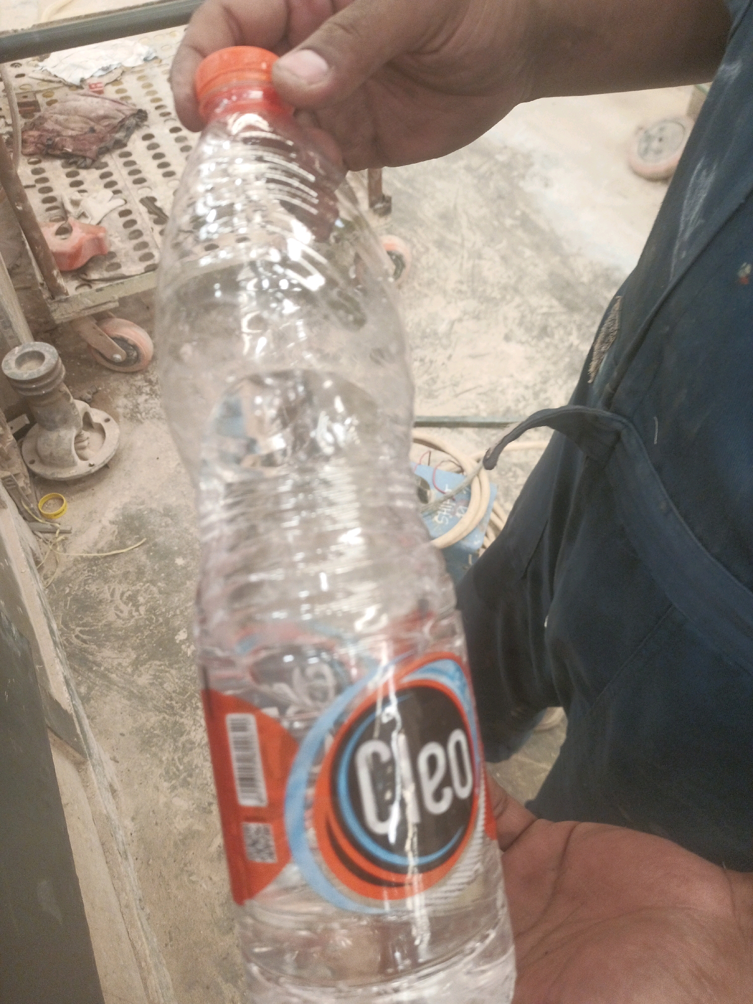 A close-up, eye-level shot captures a person's hands holding a partially crushed, clear plastic water bottle with an orange cap. The bottle has a label with the word "Cleo" in white text within a circular logo. The background appears to be a dusty, concrete floor with scattered tools and debris, including a few pink wheels and a metal fixture. A dark blue garment, possibly overalls or a uniform, is visible on the right side of the image. The setting suggests an outdoor or workshop environment in a place like Lebaniwaras, Indonesia, with evidence of construction or repair work.