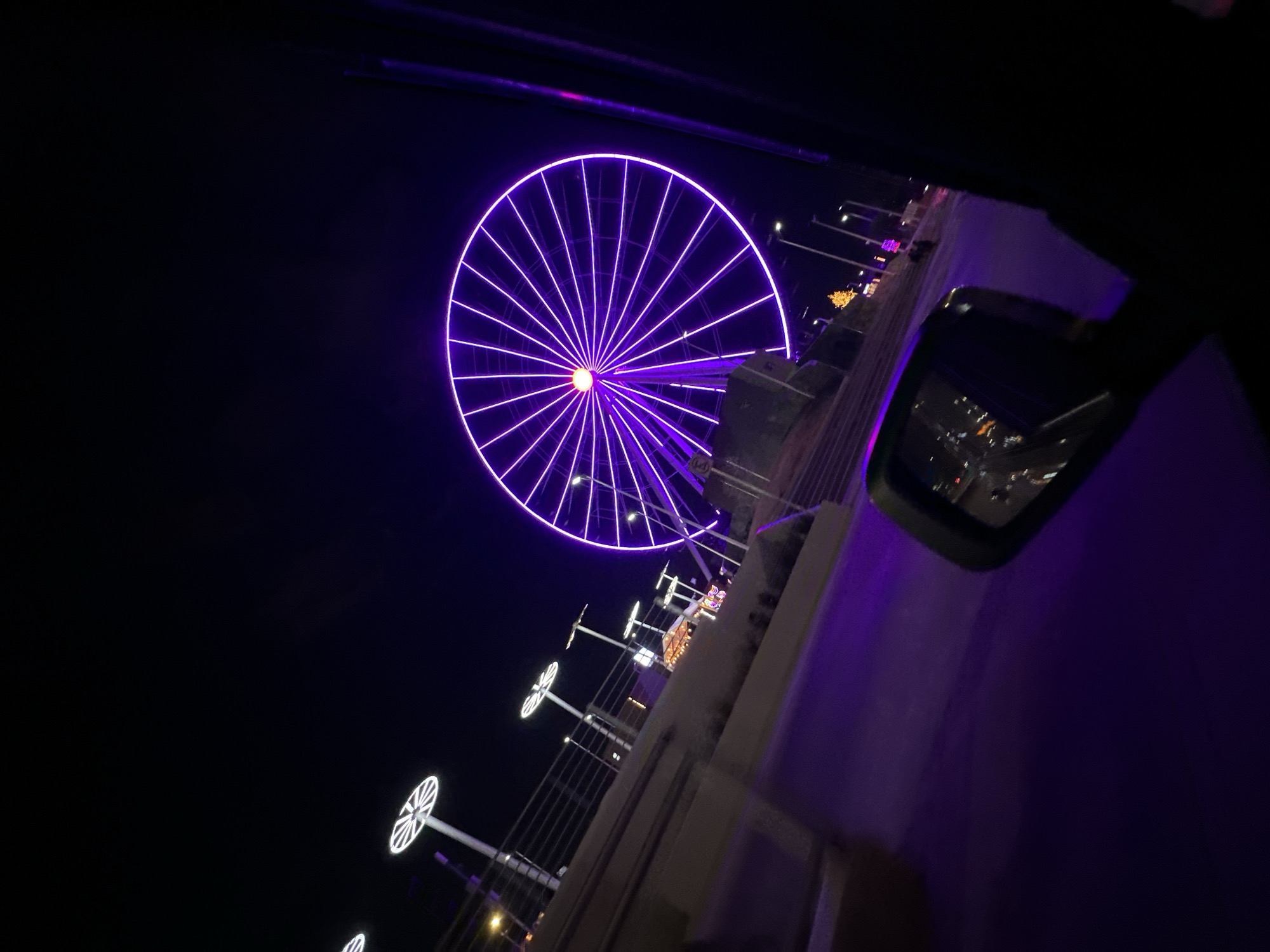 The image depicts an outdoor night scene captured from the interior of a vehicle, looking towards the left and slightly upwards. The primary subject is a large Ferris wheel, identified as the Estrella de Puebla, located in Puebla, Mexico. This Ferris wheel is prominently illuminated with vibrant purple LED lights outlining its spokes and rim, with an orange-yellow light at its central hub.
In the immediate foreground on the right, the dark A-pillar and the exterior rearview mirror of the vehicle are visible. The mirror reflects ambient lights from the street and the purple glow. To the left of the Ferris wheel, a paved surface or road reflects the purple illumination. Adjacent to this, a low concrete barrier runs parallel to the field of view, topped by a metal railing. Beyond the barrier, several modern streetlights with distinctive circular, white-lit tops extend into the midground. Further into the midground on the left, several smaller, warmly lit structures or tents are discernable. The sky above is dark and clear. No individuals are visible within the scene.