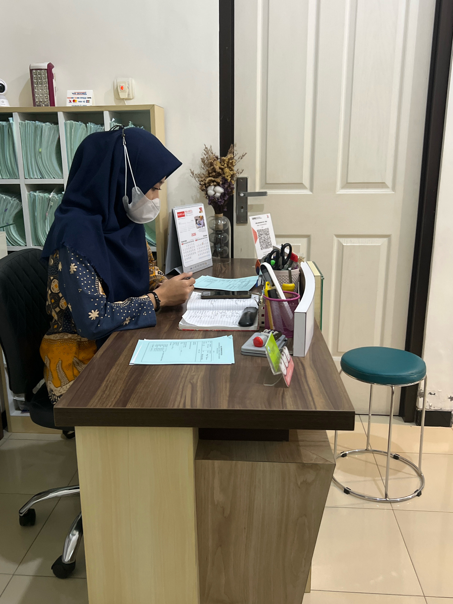 The image depicts an indoor scene, likely an office or administrative area, in Magelang, Indonesia. A woman wearing a navy blue hijab and a white face mask is seated at a wooden desk, interacting with her mobile phone. Her attire includes a batik-patterned top. The desk is cluttered with office supplies such as papers, pens, scissors, a stapler, a small calendar, and a mobile phone. Behind the woman, a shelf unit is filled with files. To the right of the desk, a white door with paneling is visible, and a teal stool with a circular chrome base is placed nearby. The flooring is tiled. The overall impression is a functional workspace.
