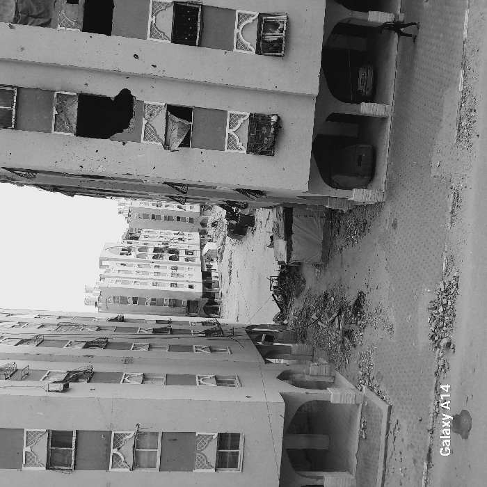 Black and white photo shows buildings on both sides of a street with rubble and debris. The buildings show signs of damage, with missing windows and parts of walls. A figure is walking in an arched walkway of a building on the right. Other structures and tents are visible amidst the debris. More buildings are in the background.