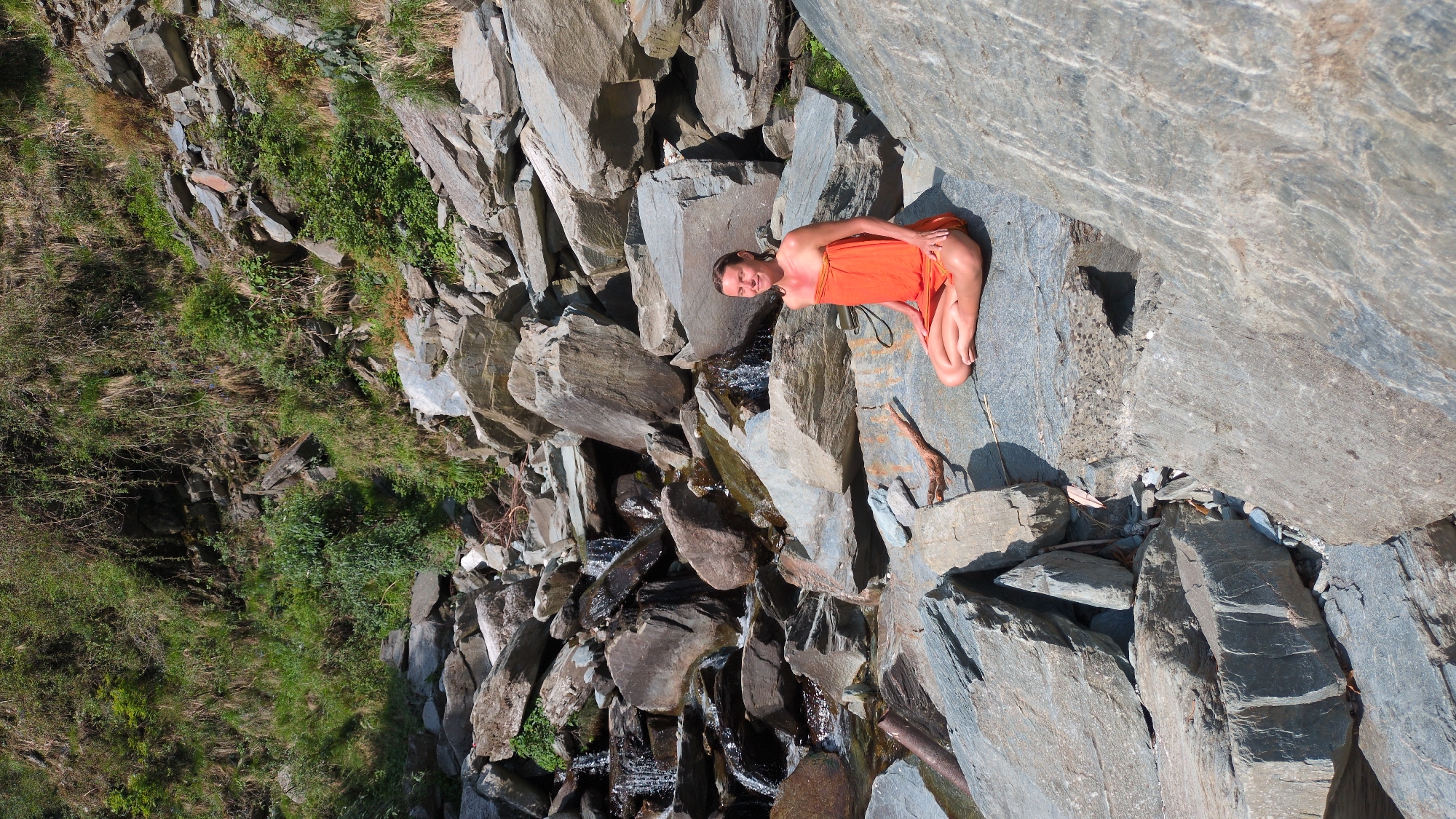 A woman in an orange garment is seated on a large, gray rock in a landscape composed primarily of similar gray rocks and greenery. A small stream or waterfall flows through the rocks near the woman. The scene is located in Bhagsu, India. The woman is looking at the camera and smiling.
