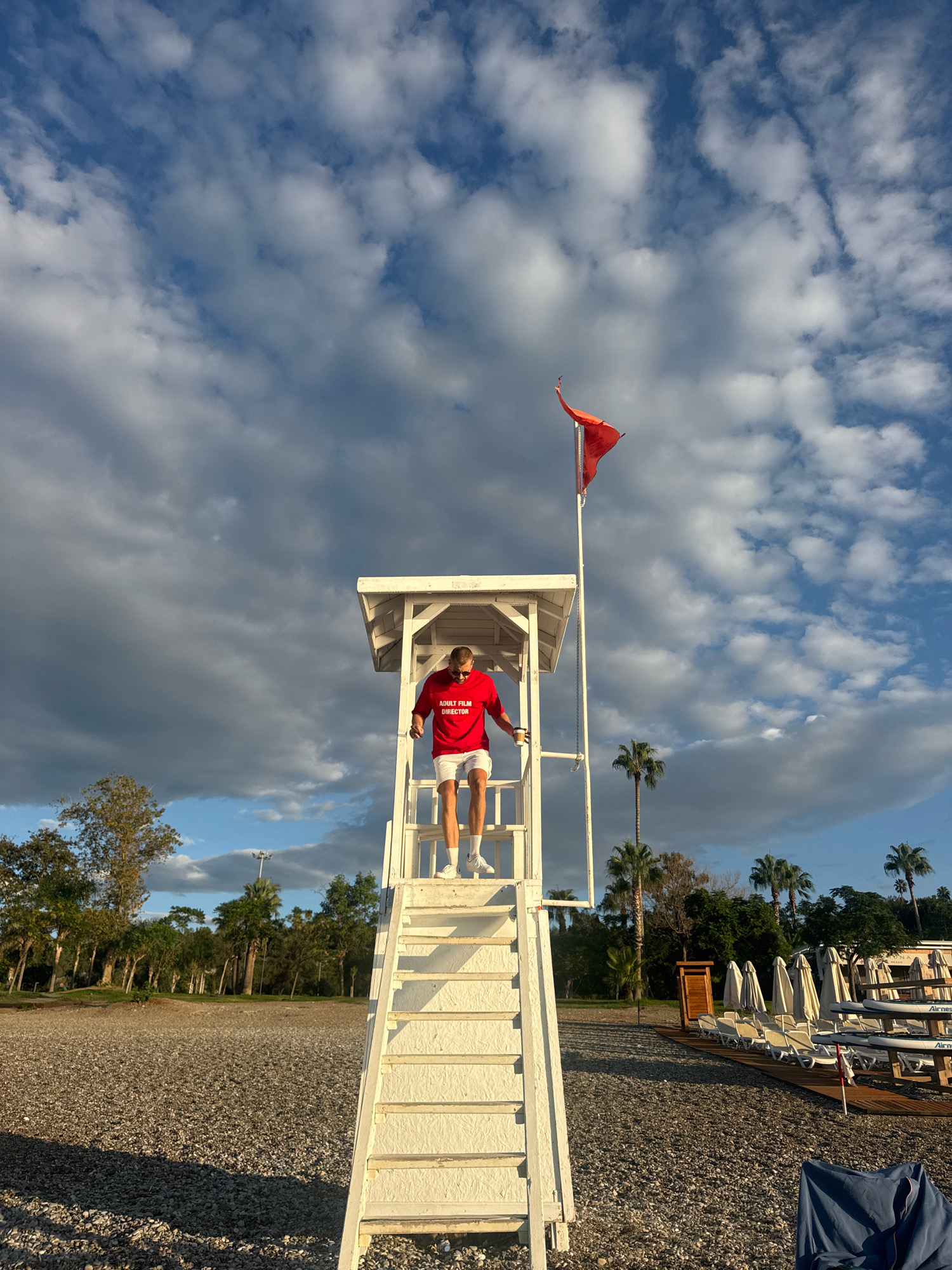A man in a red shirt and white shorts is standing on the stairs of a white lifeguard tower on a beach. He is looking down and appears to be walking down the stairs. The lifeguard tower has a red flag flying from the top. The beach is covered in pebbles and there are palm trees and lounge chairs in the background. The sky is blue with some clouds. The scene is likely in Antalya, Turkey, given the presence of palm trees and the general coastal environment.