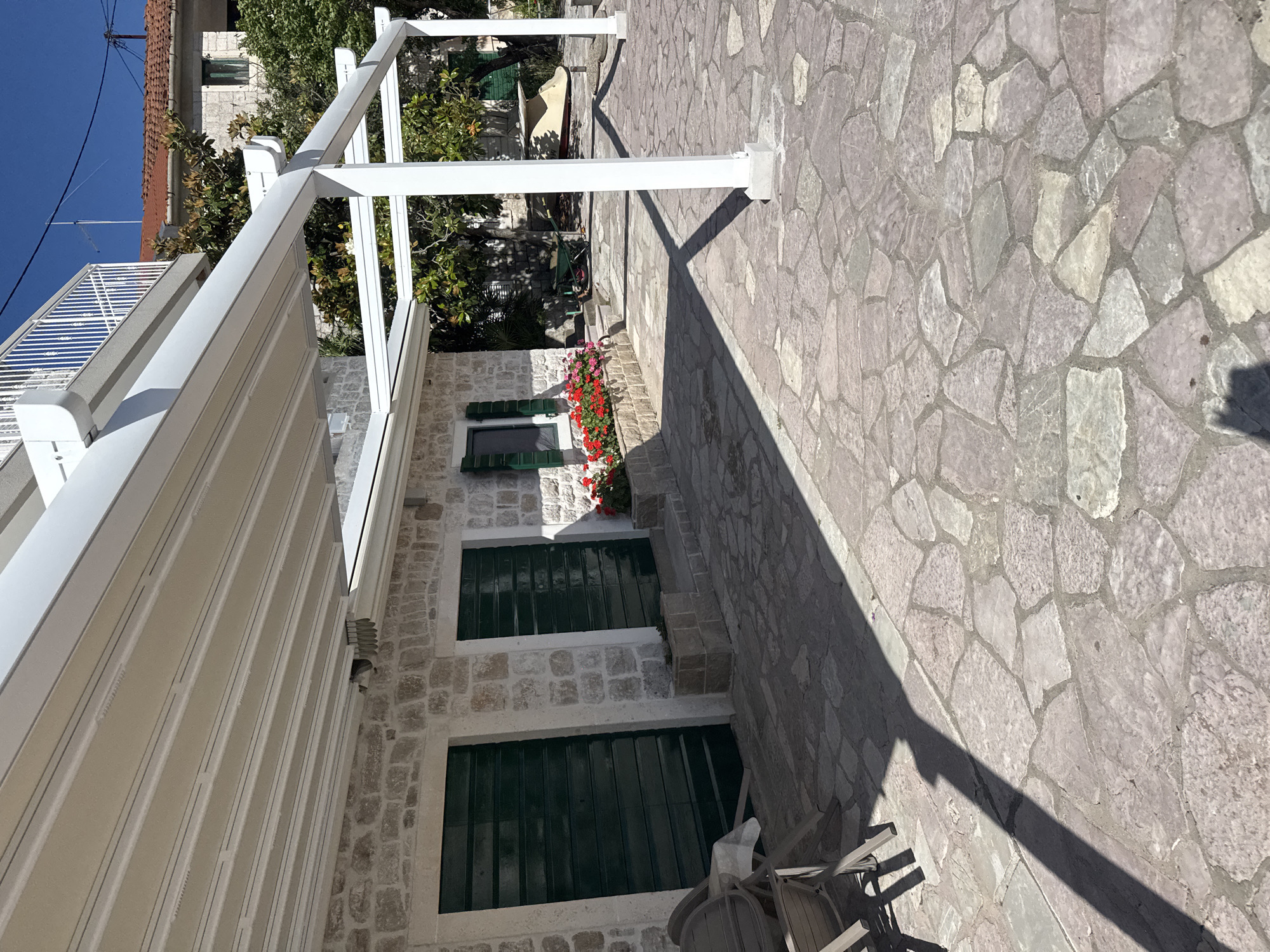 An outdoor scene in Unknown, Unknown, depicts a traditional stone building with a modern white metal pergola and retractable awning system. The building is constructed of irregular light-colored stone blocks and features two large dark green wooden doors or shutters on the left. A window with dark green louvered shutters, slightly open, is visible further right, above a stone planter box filled with red and pink flowers. Above the pergola, a white balustrade of a balcony is visible. The pergola, painted white, consists of multiple vertical posts anchored to concrete bases on the patio, supporting horizontal beams and a beige retractable fabric awning. The patio is paved with irregularly shaped flagstones in shades of grey, beige, and reddish-brown. Strong sunlight casts distinct shadows across the patio. In the background, green trees and another building with a red-tiled roof are present under a clear blue sky. A small green wheelbarrow and a light-colored object, possibly a boat, are visible amongst the trees. Part of a brown outdoor chair is visible in the foreground on the left.