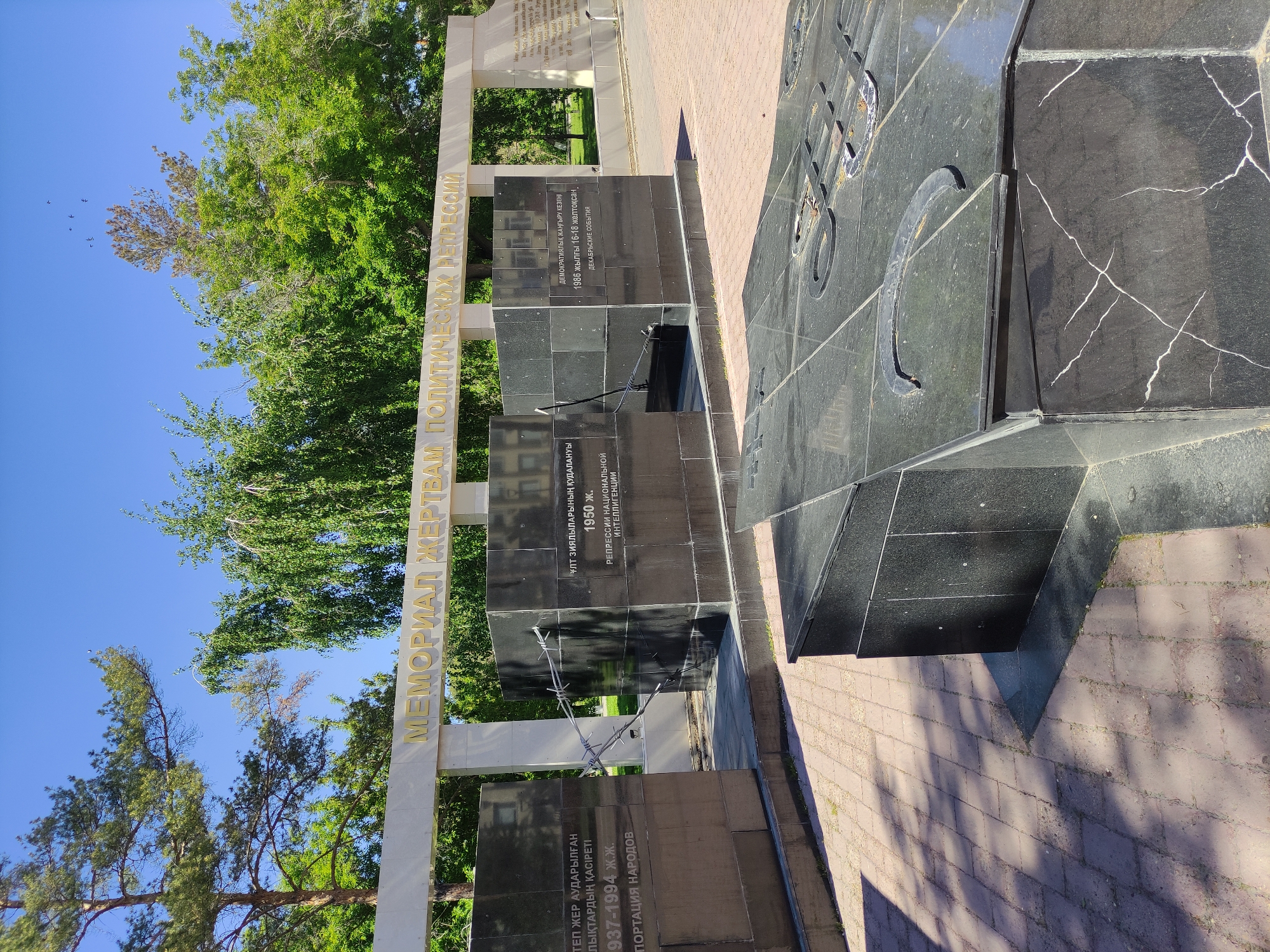 The image shows a memorial in Pavlodar, Kazakhstan, dedicated to the victims of political repression. It features a white archway with golden Cyrillic lettering spelling out "Memorial to the Victims of Political Repression." Behind the archway are several dark marble monuments with inscriptions in Kazakh and Russian, detailing different periods of repression, including the deportation of peoples (1937-1994) and the persecution of the national intelligentsia (1950). There is barbed wire draped over one of the monuments. To the right is a large block of dark marble with what appears to be a Soviet emblem etched on its surface. The ground is paved with red bricks, and trees are visible in the background, suggesting it is located in a park or public space. The sky is blue and clear.