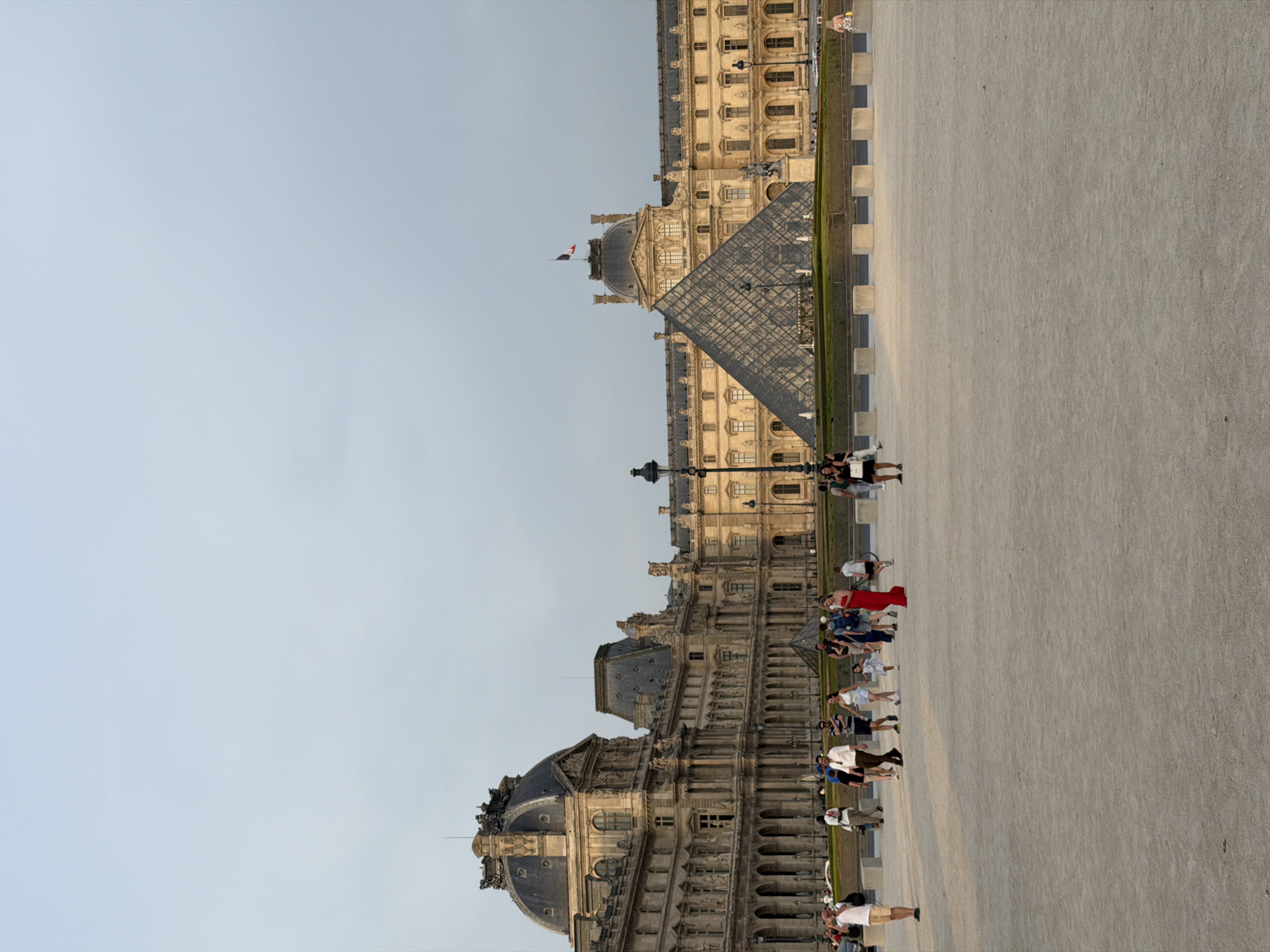 The image depicts the Louvre Museum in Paris, France, a renowned cultural institution with distinctive architecture. The foreground is an open, paved area, possibly a plaza or courtyard, with a light-colored gravel surface. A group of tourists are visible in the middle ground, walking and interacting. Among them, a woman in a vibrant red dress stands out. The background features the iconic Louvre Palace, a grand building with classical architectural elements, including ornate facades, arched windows, and domes. The prominent glass pyramid, a modern addition designed by I.M. Pei, is situated in front of the palace, reflecting the sky and surrounding structures. The sky above is a pale, overcast blue. The lighting suggests it is late afternoon or early evening, with soft shadows cast across the scene. The overall atmosphere is one of a bustling tourist attraction.