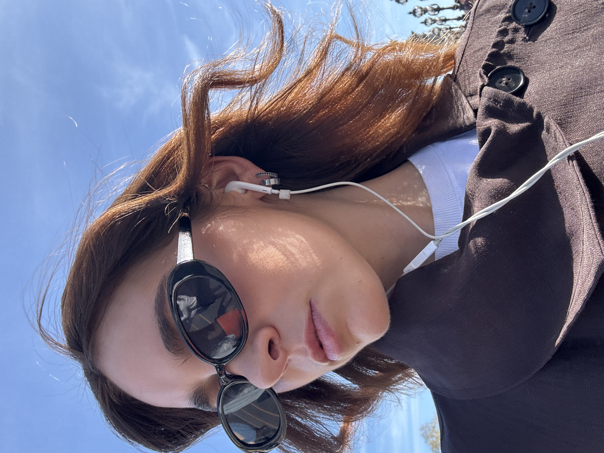 A tilted low-angle shot focuses on a woman with light skin, brown hair, and wearing sunglasses, listening to earphones, and wearing a brown jacket. The background reveals a clear blue sky. The reflection in the sunglasses shows the Eiffel Tower, indicating the location as Paris, France. The woman is wearing a white shirt and has metal rings in her ear. The brown jacket has two black buttons. The composition is asymmetrical and captures a candid moment.