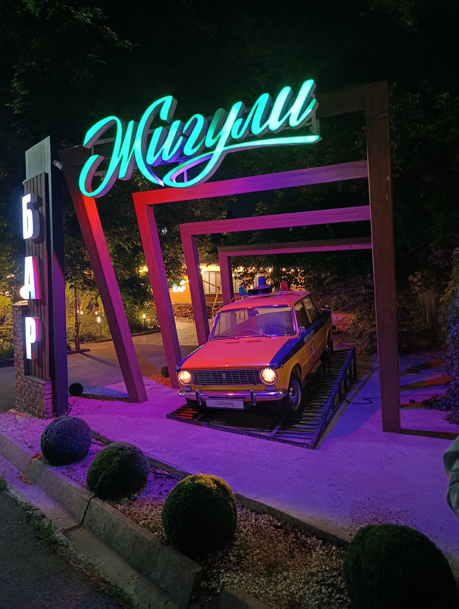 The image shows the entrance to a bar named "Zhiguli" (likely after the car brand) in Almaty, Kazakhstan. A vintage orange and blue Zhiguli car, resembling a police vehicle, is displayed on a platform within a wooden frame structure. Above the car is a neon sign spelling "Zhiguli" in a cursive script. To the left of the frame is a vertical sign spelling "БАР" (BAR) in Cyrillic letters. Three round bushes are planted along the sidewalk leading to the entrance. The lighting is colorful, with pink and purple hues illuminating the scene. The setting appears to be nighttime. In the background, there is a glimpse of a building with illuminated windows, and outdoor seating can be seen.