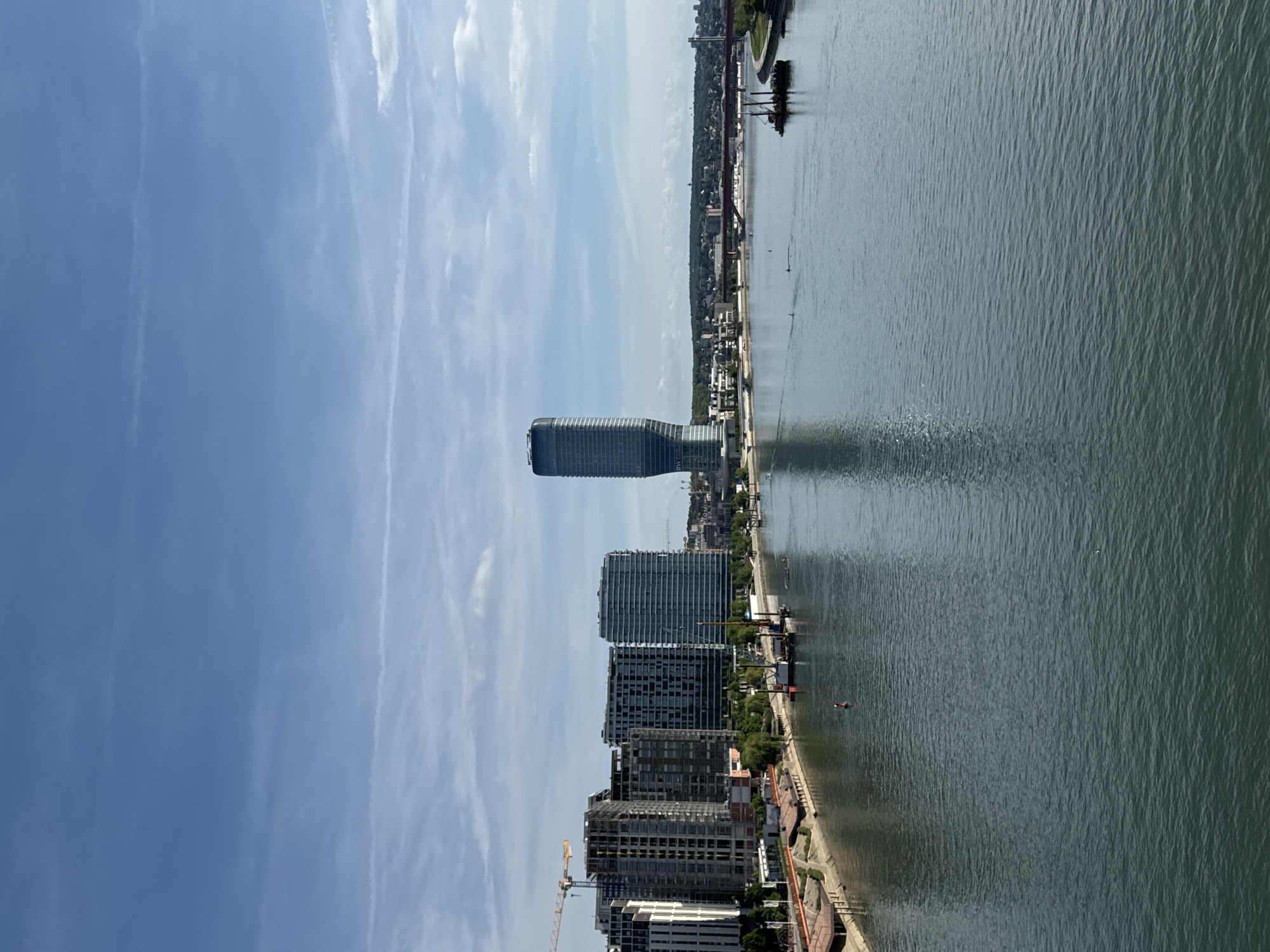The image captures a daytime view of Belgrade, Serbia, featuring the Sava River and the Belgrade Waterfront development. The skyline is dominated by the Belgrade Tower, a modern skyscraper with a distinctive design. Several other high-rise buildings, some still under construction, are visible along the riverbank. A construction crane is also present in the image. The Sava River stretches towards the horizon, reflecting the sky and the buildings on the shoreline. A bridge is visible on the horizon over the river. The sky is blue with scattered clouds. The image is taken from an elevated perspective.