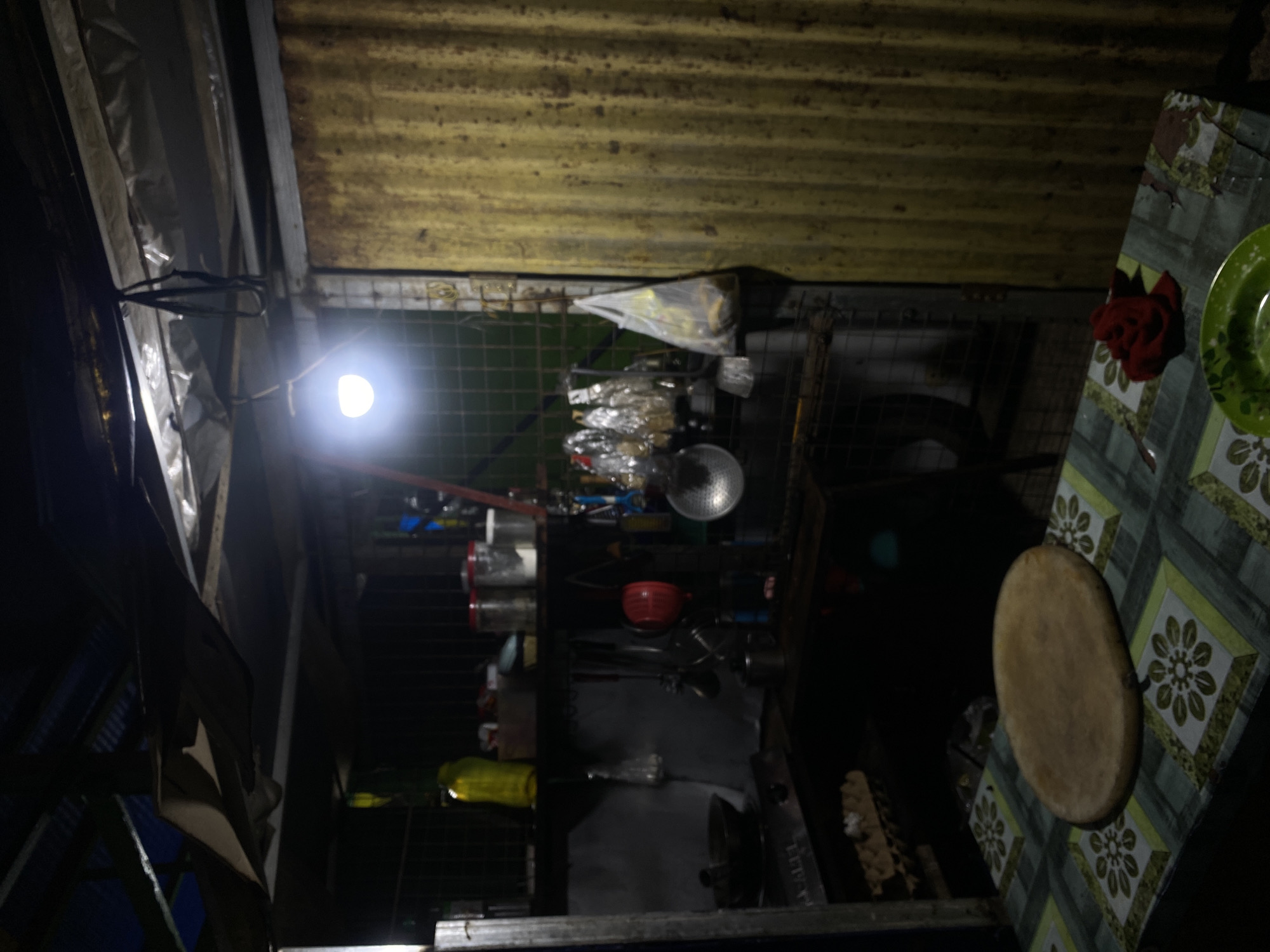 The image depicts an outdoor makeshift kitchen in a dimly lit environment. The scene is characterized by a corrugated yellow metal wall on the right, partially covering the background. A metal shelf unit is visible, holding various kitchen items including jars, bottles, and utensils. A bright LED bulb hangs from the ceiling, illuminating the space. In the foreground, a table covered with a patterned cloth displays a round wooden cutting board, a red cloth, and a green bowl. The overall impression is one of a humble, functional kitchen space, likely located in a residential area within Davao City, Philippines. The presence of basic cooking implements and the rustic construction suggest a practical, everyday setting.
