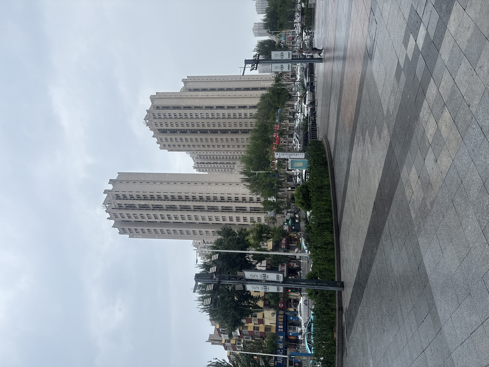 This image depicts a street scene in Qinhuangdao, China. In the background, two large, light-colored apartment buildings with numerous windows dominate the skyline. The sky is overcast and a pale gray. In the mid-ground, a street with several cars is visible, lined with trees and smaller buildings with colorful facades. In the foreground, a wide pedestrian walkway paved with large gray tiles leads towards the street. A lamppost with multiple lights and banners stands on the left side of the walkway. Two people are walking on the walkway in the right mid-ground. The overall impression is of a typical urban environment on a cloudy day.