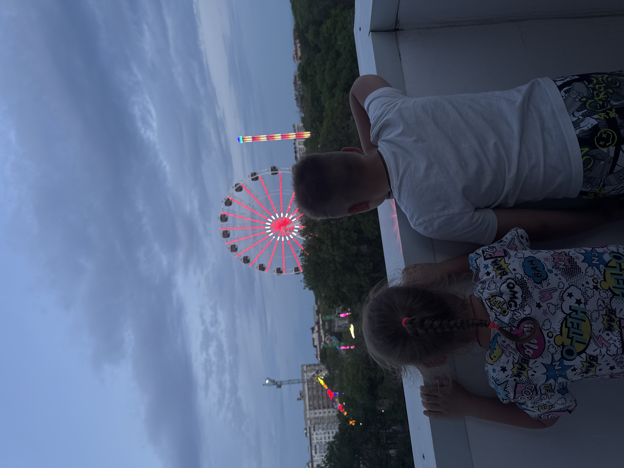 Two children, a boy and a girl, are looking over a ledge at a Ferris wheel and other attractions in what appears to be a fairground or amusement park. The boy is wearing a white t-shirt and patterned shorts, while the girl has a white t-shirt with a cartoon print and her hair is in a braid with pink ties. The Ferris wheel is lit up with red lights. In the background, trees and buildings are visible. The sky is a dusky blue. This image shows the city of Yevpatoriya in Ukraine.