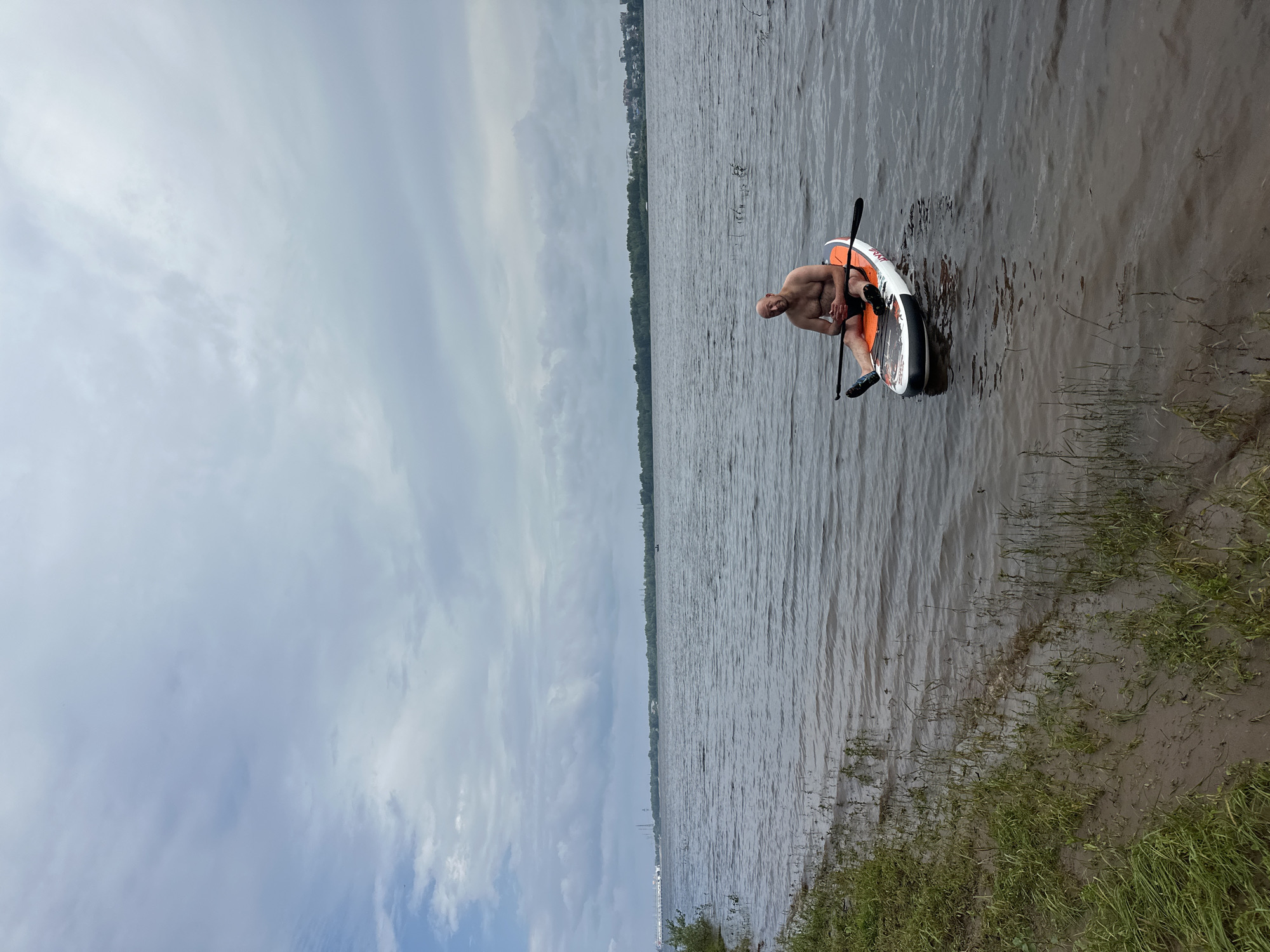 A bald man with a tan torso and orange swim trunks sits on a white and orange paddleboard in a wide river. He holds a black paddle across his lap. The river is calm with small ripples. To the left of the frame, a grassy bank slopes down to the water. In the distance, a tree-lined shore stretches across the horizon, with a few buildings visible on the far right. The sky is overcast with grey clouds. The man is positioned slightly to the right of the center of the image.