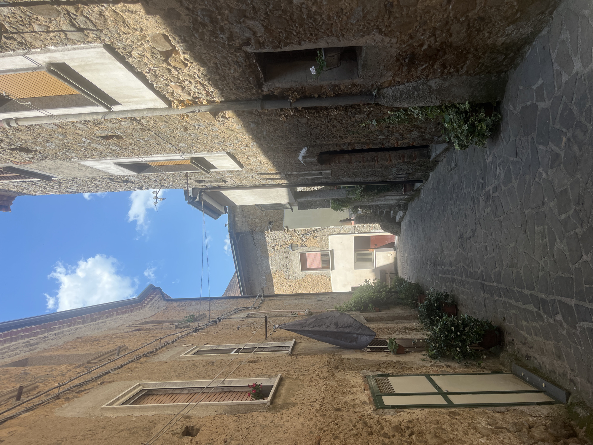 This is a ground-level, eye-level, medium shot of a narrow cobblestone alleyway in Vallo della Lucania, Italy. The alley is flanked by tall, aged buildings constructed from rough-hewn stone and stucco. The left side of the alley features a building with a weathered, textured facade, a window with brown shutters, and a smaller window above it. A dark, flag-like object hangs from a line attached to the building. Another window with green trim is visible lower down. Lush green plants in pots are placed along the base of this building.
The right side of the alley is dominated by a stone building with a textured, uneven surface. It has several windows with light-colored frames and dark shutters, one of which is partially open. A vertical pipe runs down the facade of this building. A small window opening near the bottom of this building contains a planter with white flowers.
The alleyway itself is paved with irregularly shaped grey cobblestones. Sunlight casts shadows across the alley, indicating it's daytime. The sky above is bright blue with scattered white clouds. The scene suggests a quiet, historic part of town, with architectural details indicative of Mediterranean or southern European building styles. There are no people visible in the image. The overall impression is one of a charming, somewhat secluded European street.