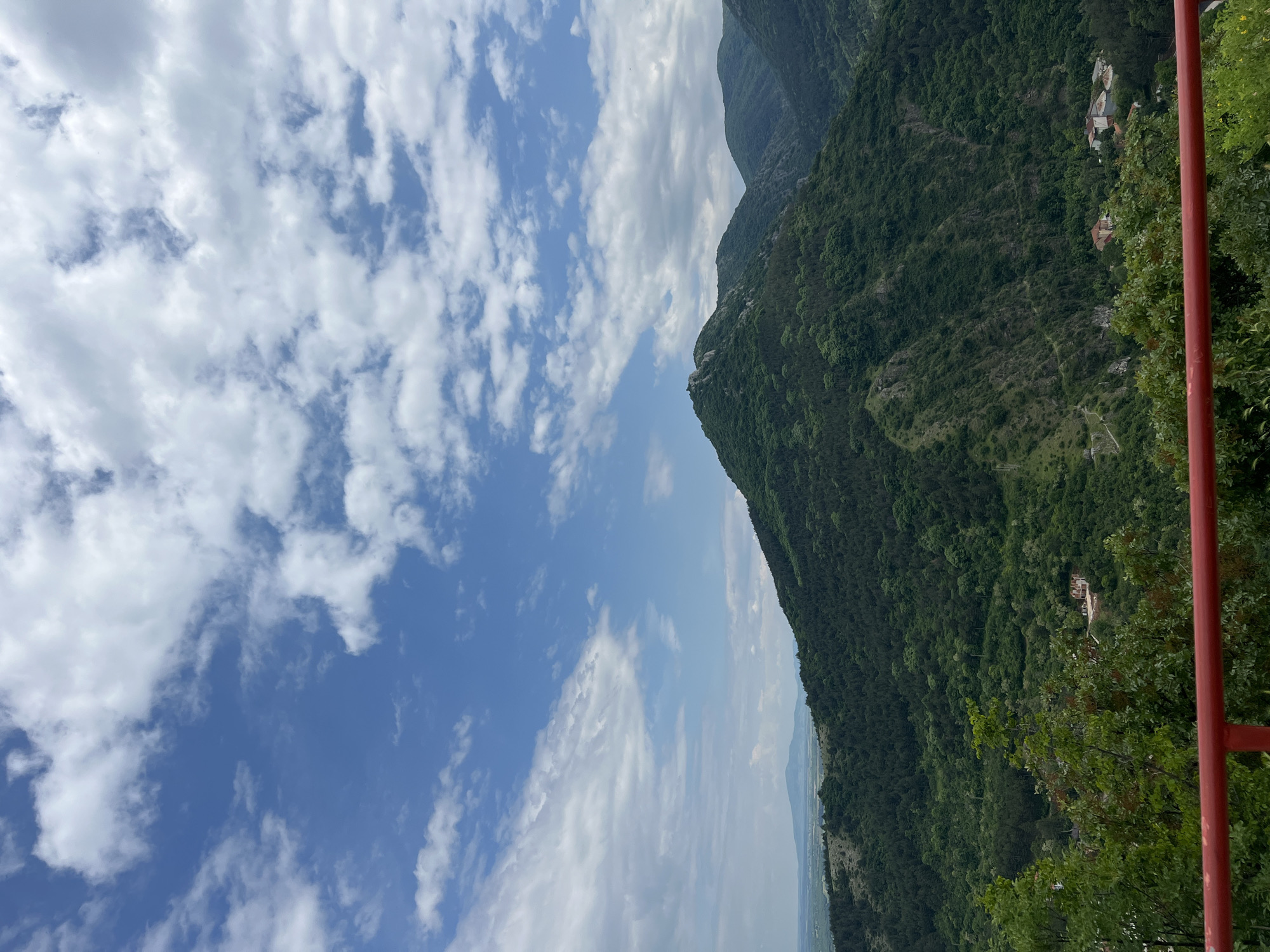 The image shows a mountain landscape in Asenovgrad, Bulgaria. The mountain is covered in green trees and vegetation. The sky is blue with white clouds. A red railing is visible on the right side of the image. The picture appears to be taken from an elevated vantage point.
