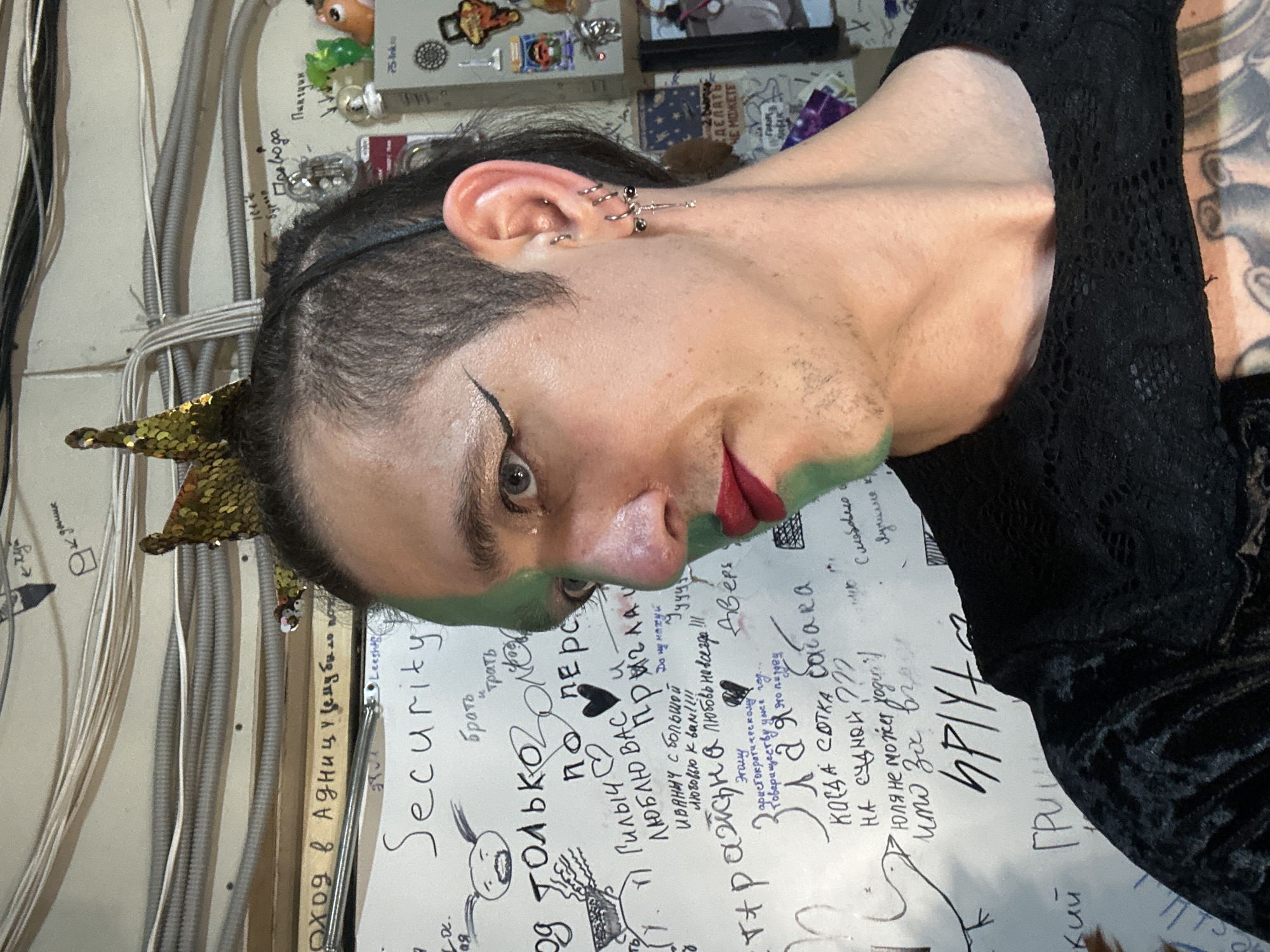 This portrait, captured in Saratov, Russia, features a person with dark hair styled with shaved sides and a longer top, adorned with a golden sequined crown. They wear expressive makeup, including dramatic black eyeliner, red lipstick, and green face paint around the lower cheeks. The visible ear is heavily pierced with multiple rings and a prominent dangling charm. A black lace top is visible, along with a tattooed arm on the right side of the frame. The person gazes directly at the camera with an intense or serious expression, presenting an artistic and self-styled image.
The background reveals an indoor setting with a light-colored wall featuring exposed electrical wiring, some neatly bundled and others running through corrugated conduits. Above these wires, small decorative items and stickers are affixed, including a small animal figurine. Below the wiring, a paper-covered section of the wall is filled with handwritten Russian text and doodles. Prominent phrases include "Security," "Оход в Адницу" (Entry to Hell), "Провода" (Wires), "СДЕЛАТЬ ЧЕ МОЖЕТЕ" (DO WHAT YOU CAN), "Гилыч ЛЮБЛЮ ВАС" (Gilych I LOVE YOU), "Иваныч с большой Любовью к вам!!!!" (Ivanych with great Love to you!!!!), and "ПРИВЕТ" (HELLO). Other personal or colloquial messages are also present, contributing to a casual, personalized, and possibly creative environment. The image depicts a moment of self-expression within a distinct, informal space.