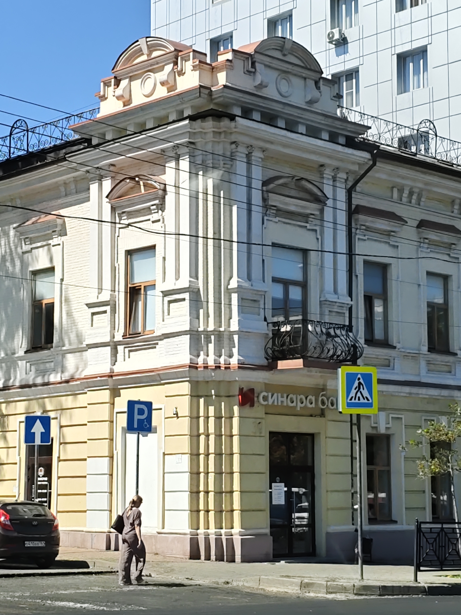 The image shows a street view in Rostov-on-Don, Russia, featuring an ornate cream-colored building with yellow accents. The building has decorative cornices, columns, and arched windows, with a wrought-iron balcony on the second floor. A modern white building with a grid facade is visible behind it. In the foreground, a person in a grey jumpsuit walks away from the camera on the sidewalk. A dark car is parked on the left. Two street signs are visible: one with an upward arrow and another with a "P" symbol indicating parking and a wheelchair accessibility icon. The building has the name "Синара Банк" (Sinara Bank) on its facade, suggesting it is a bank. The scene is bathed in sunlight, casting shadows.