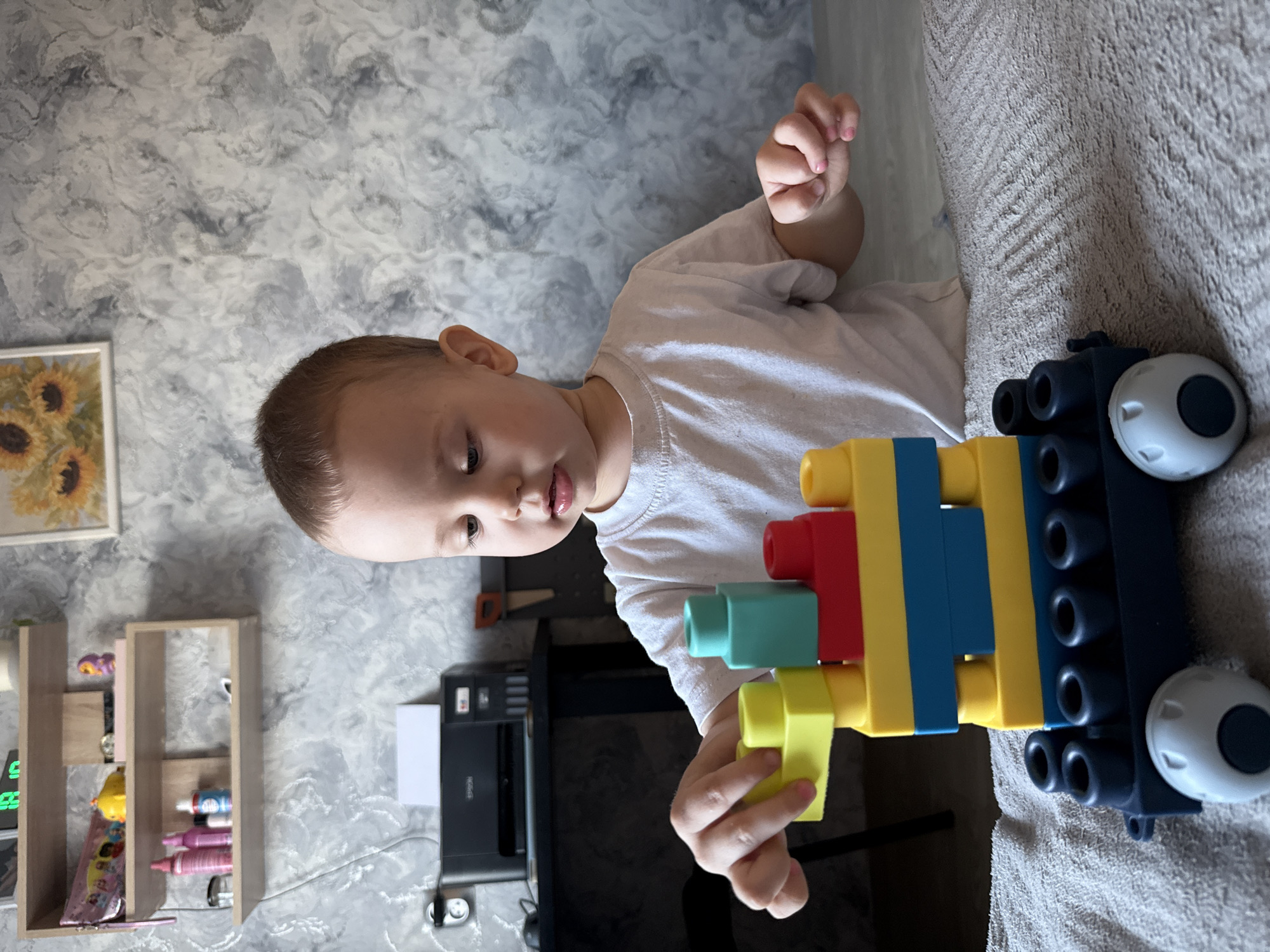 A young child is playing with large building blocks on a gray surface. The blocks are various colors including yellow, blue, red, and teal, and are assembled into a car shape. The child is wearing a white t-shirt. In the background, a printer sits on a table along with a shelf displaying small decorative items, and a framed picture of sunflowers hangs on the wall. The location of the scene is indoors, at an unknown location.