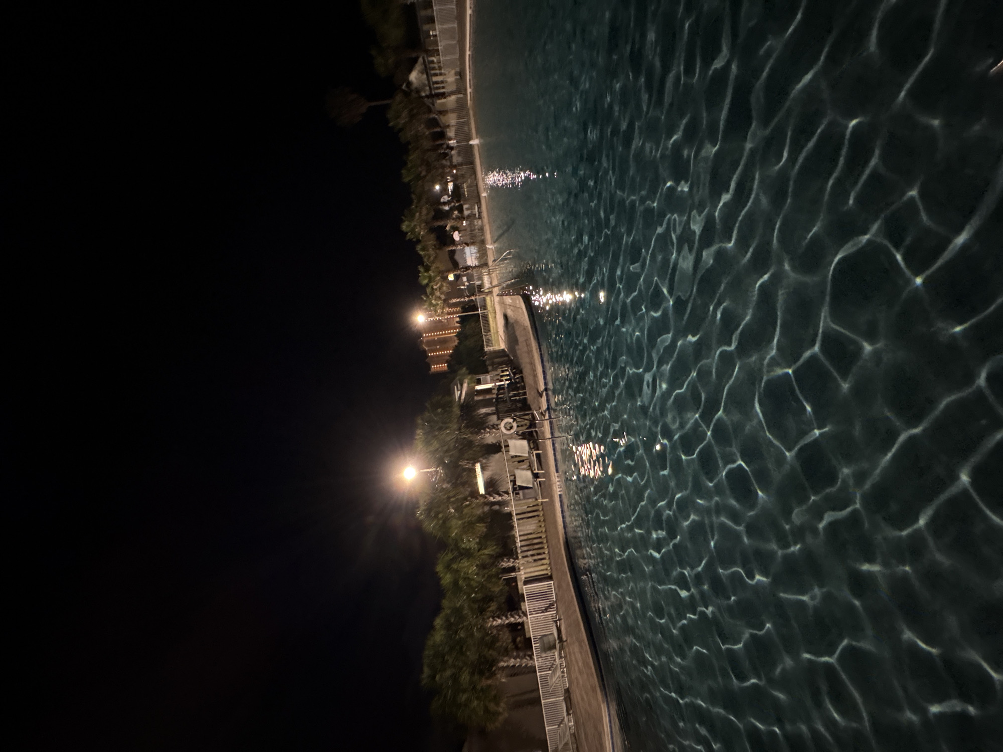 The image shows a swimming pool at night in Panama City Beach, United States of America. The pool is illuminated by artificial lights, creating reflections on the water's surface. A dark sky is visible, contrasting with the illuminated pool area. The pool is bordered by a paved walkway with palm trees and other landscaping. There are lounge chairs and tables, indicating a resort or recreational setting. A life preserver is visible.