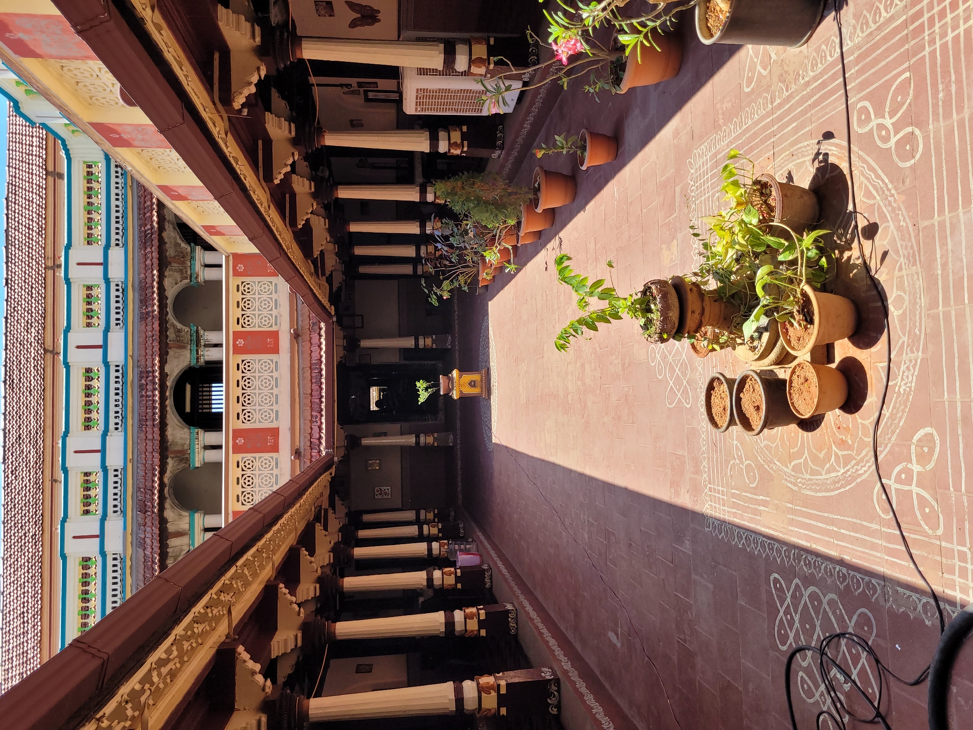 This media file captures a long, traditional Indian-style courtyard or hallway, viewed from an elevated perspective looking along its length.
On the left side, a covered walkway or verandah runs parallel to the viewer, supported by a series of white, fluted pillars with ornate dark bases and capitals. The roof structure above this walkway is visible, featuring intricate wooden elements and colorful painted sections in blue, yellow, red, and white, adorned with geometric patterns. Beyond the pillars, the interior appears darker, with glimpses of furniture, a modern air cooler, and a decorative butterfly cutout on a wall.
The right side of the image features a sun-drenched open space, paved with terracotta-colored tiles. Numerous potted plants are arranged along this tiled area, receiving direct sunlight. These include leafy green plants, some with small pink or yellow flowers, all housed in terracotta pots. White kolam (rangoli) patterns, featuring elaborate geometric and linear designs, are drawn on the tiled floor. A black electrical cable is also visible on the floor near these patterns and plants.
A strong, diagonal shadow cast by the roof structure stretches across the tiled floor, creating a clear division between light and shade, suggesting a time of day when the sun is relatively low, such as morning or late afternoon. At the far end of the courtyard, in the darker background, a small, ornate yellow or golden shrine-like object is visible.
The scene is devoid of people, conveying a sense of quietude. The architectural style, kolam patterns, and traditional elements point to a cultural setting, likely in South India.